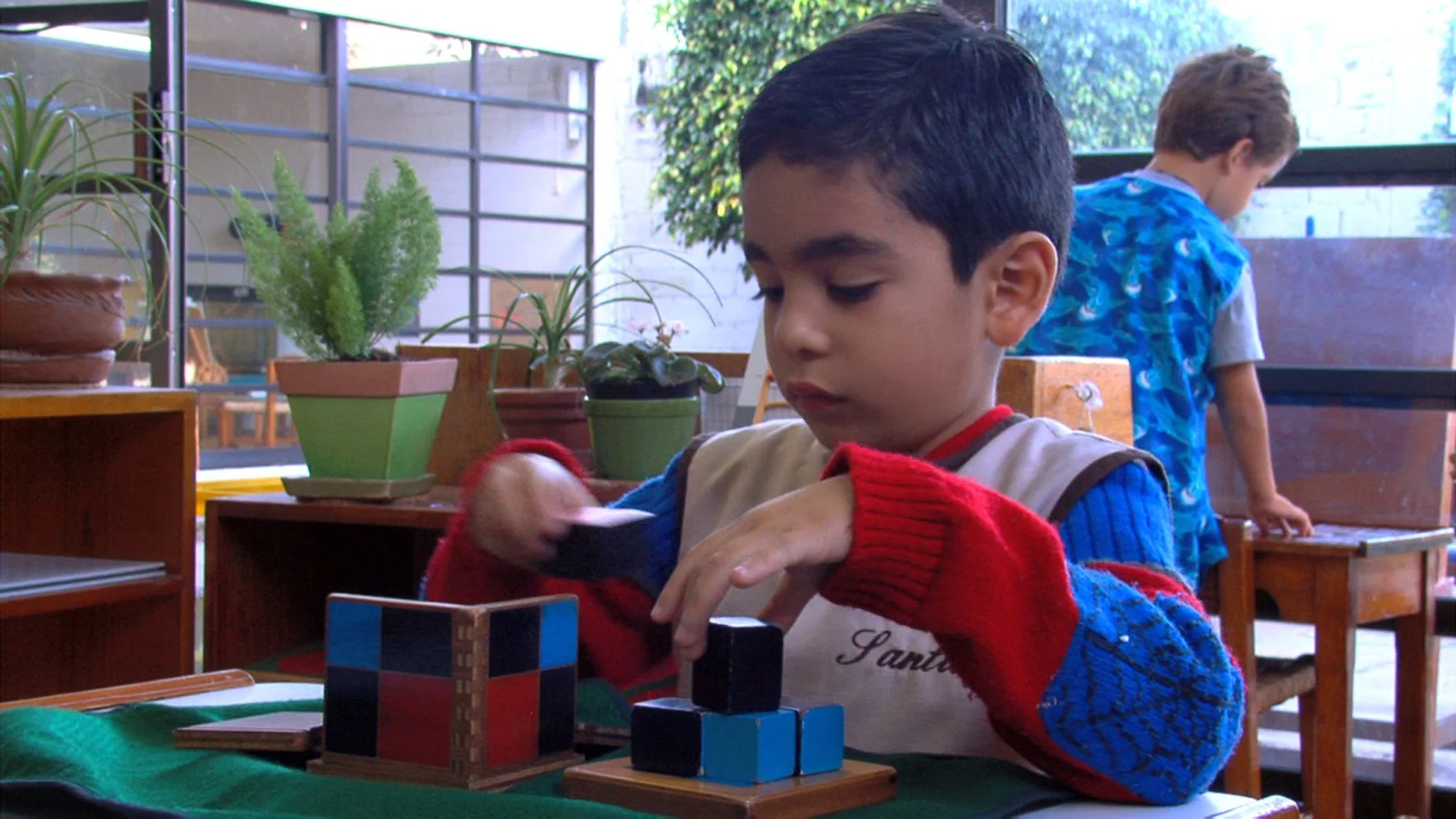A young boy playing with wooden blocks at a table inside a room with large windows and potted plants.