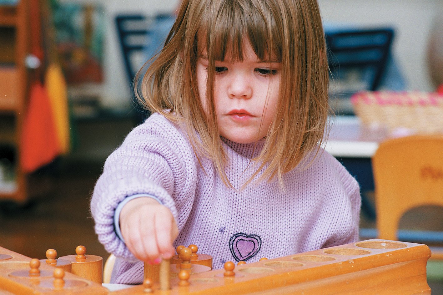 A young girl with light brown hair wearing a purple sweater playing a wooden game in a classroom.