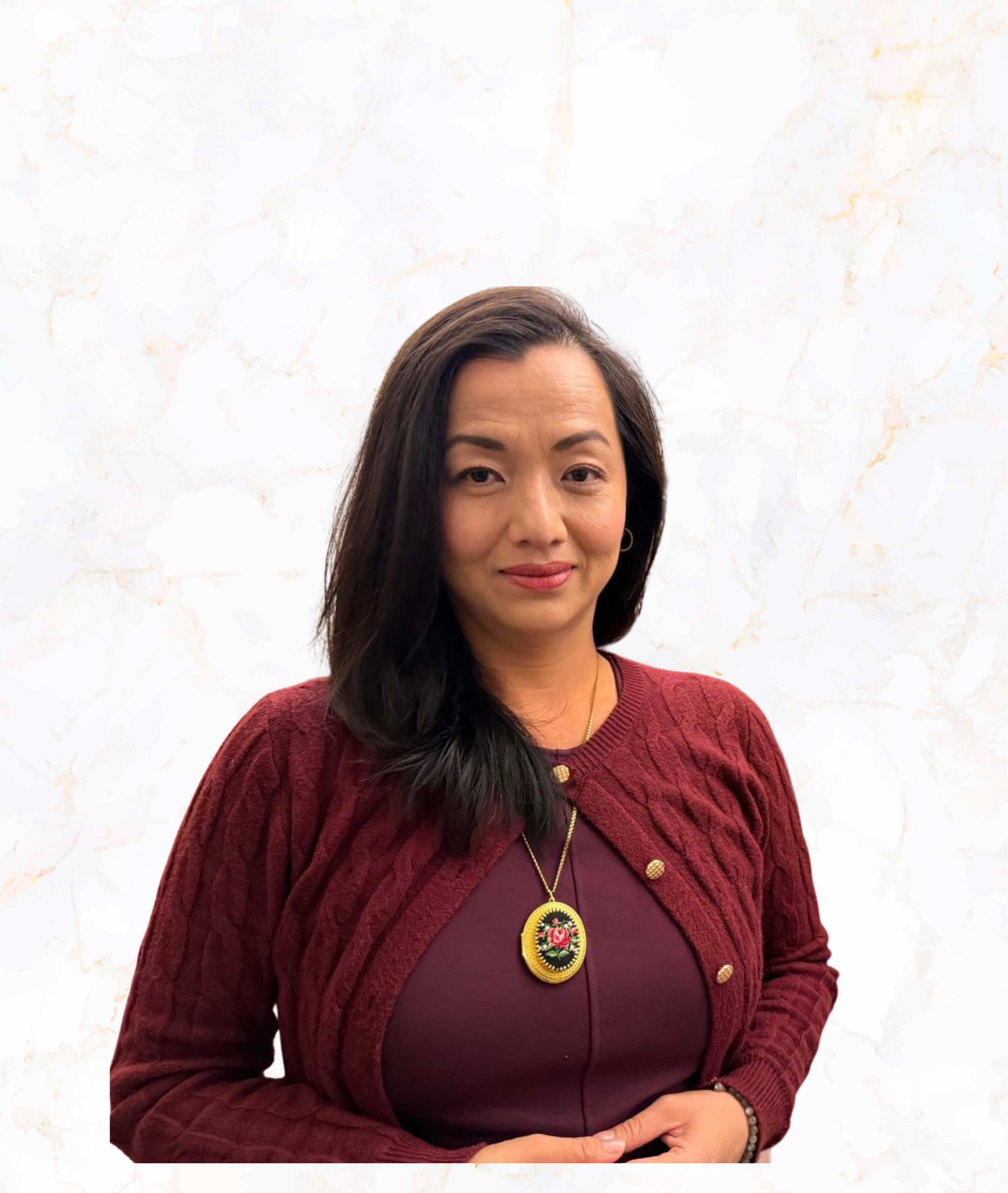 A woman with dark hair wearing a maroon sweater with a matching shirt, a gold necklace with a floral pendant, standing against a white marble background.