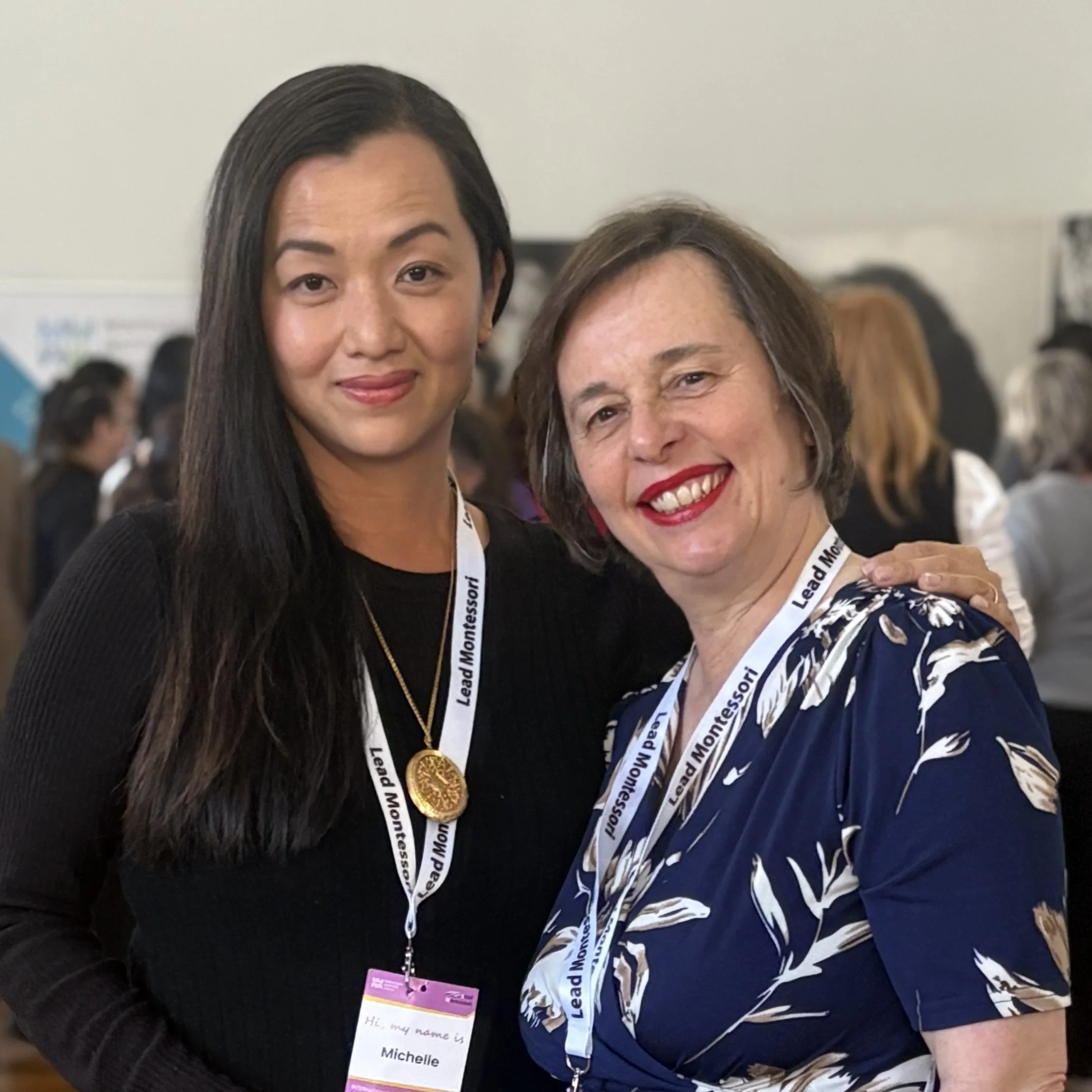 Two women smiling at a conference, wearing lanyards and badges, with a blurred background of other attendees.