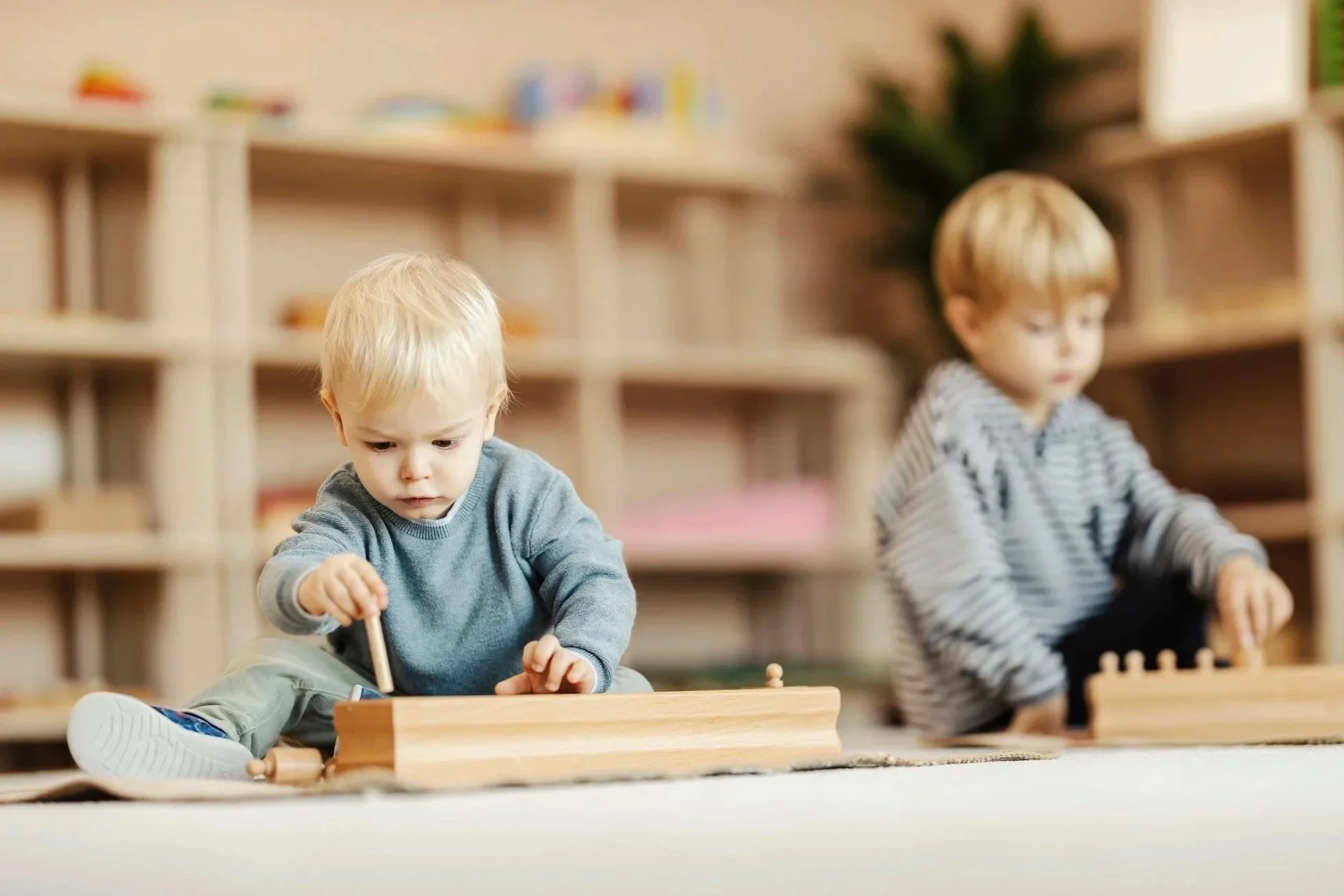 Two young children playing with wooden blocks indoors in a room with shelves and a plant.
