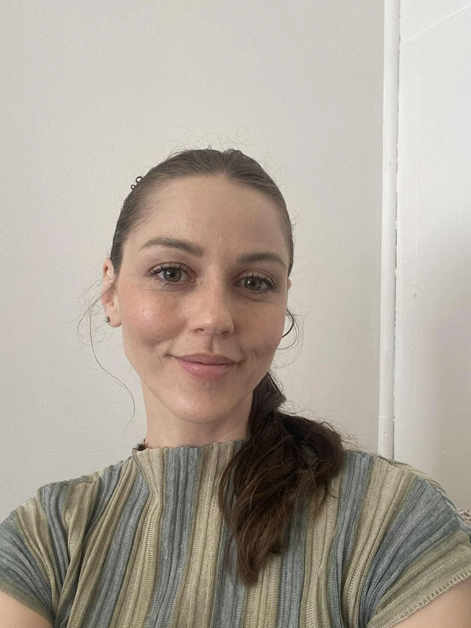 A young woman with brown hair pulled back, wearing a striped shirt, smiling in front of a plain white wall.