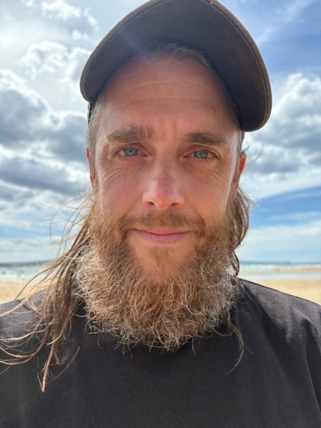 A man with a beard wearing a baseball cap, standing on a beach with the ocean and cloudy sky in the background.