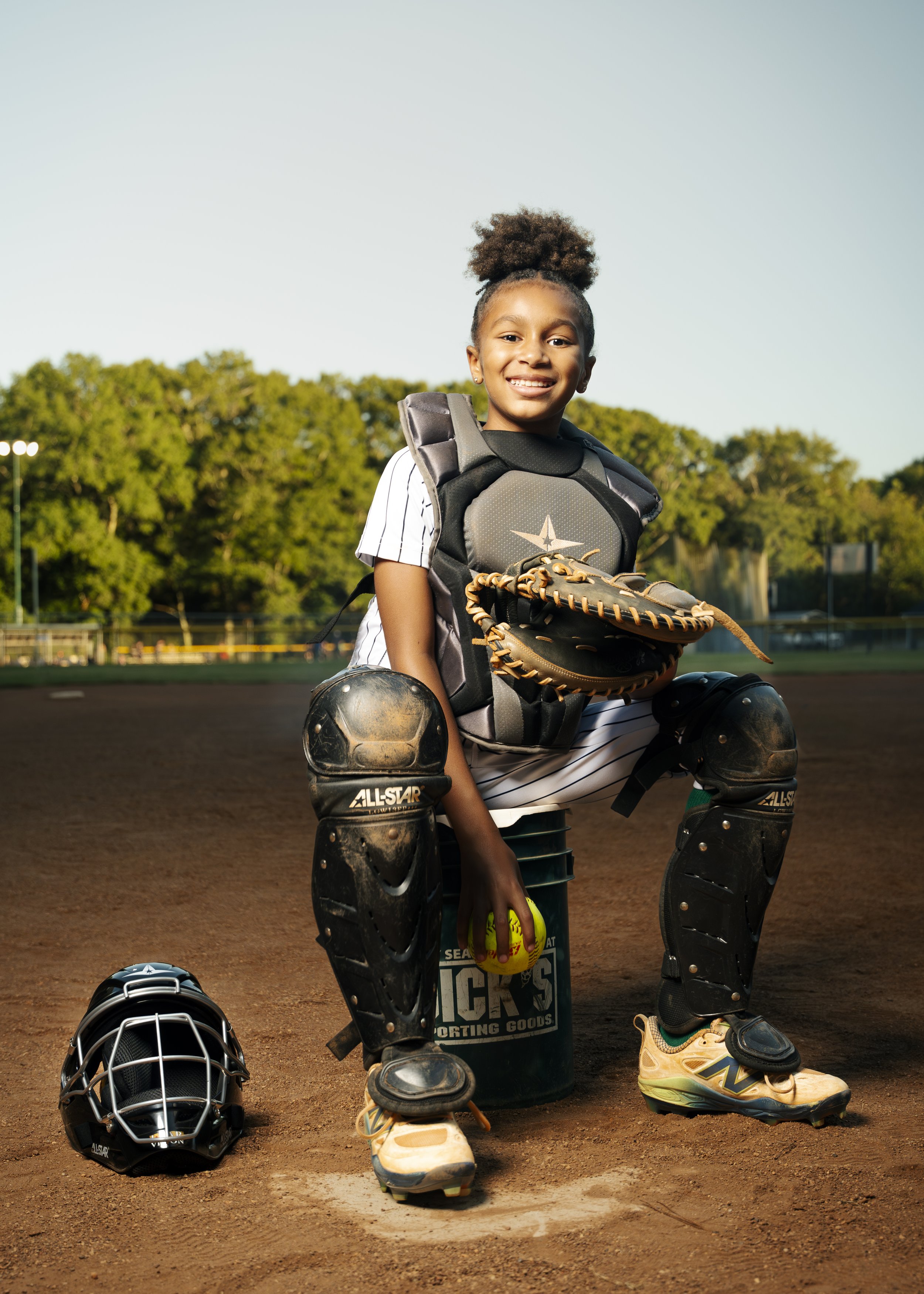 A young girl in a baseball catcher's pose on a baseball field, wearing catcher's gear including leg guards, a helmet on the ground, a bat, and holding a softball, with a baseball glove on her chest and a smiling expression.