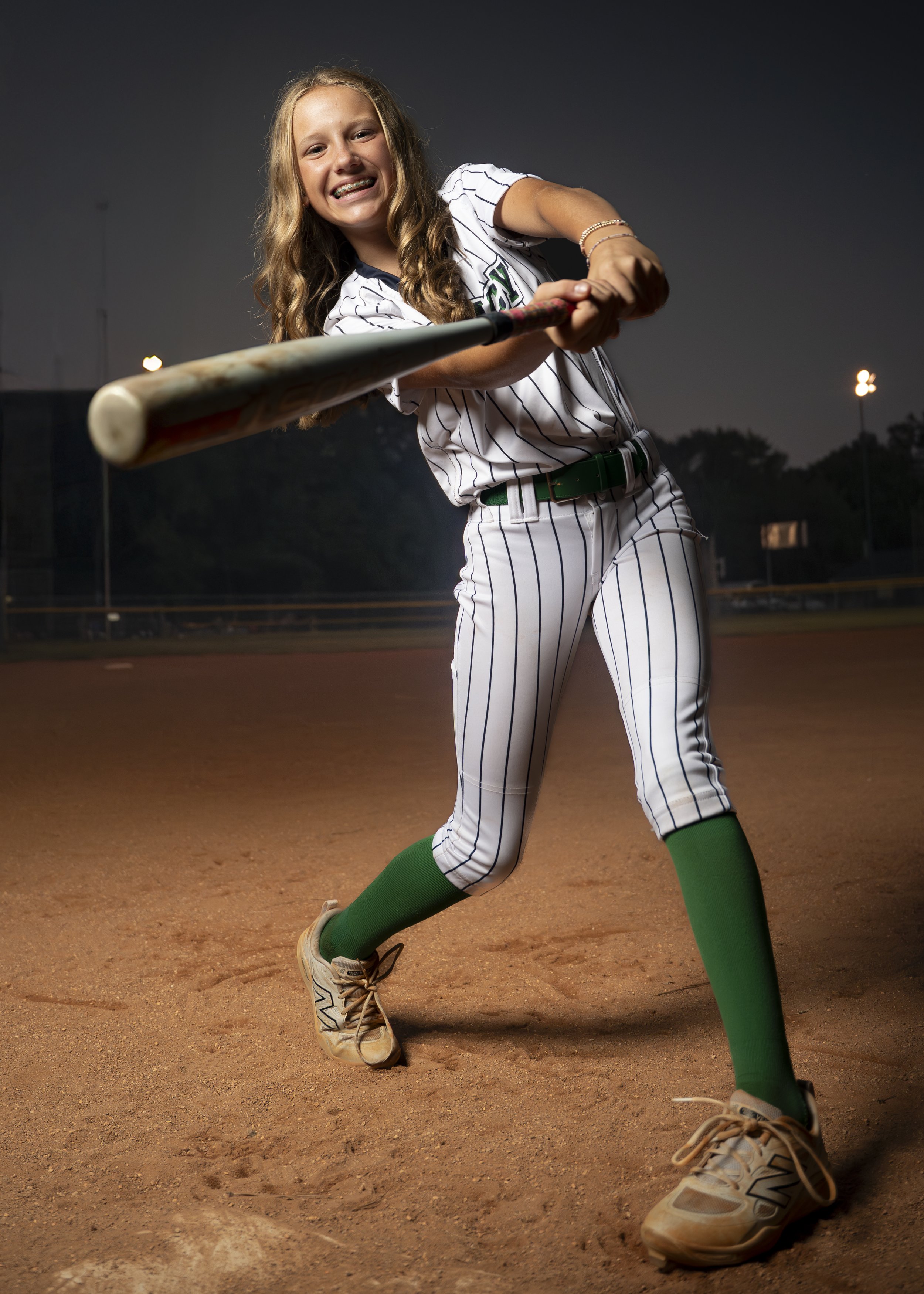 Young girl in a baseball uniform swinging a bat on a baseball field at night.