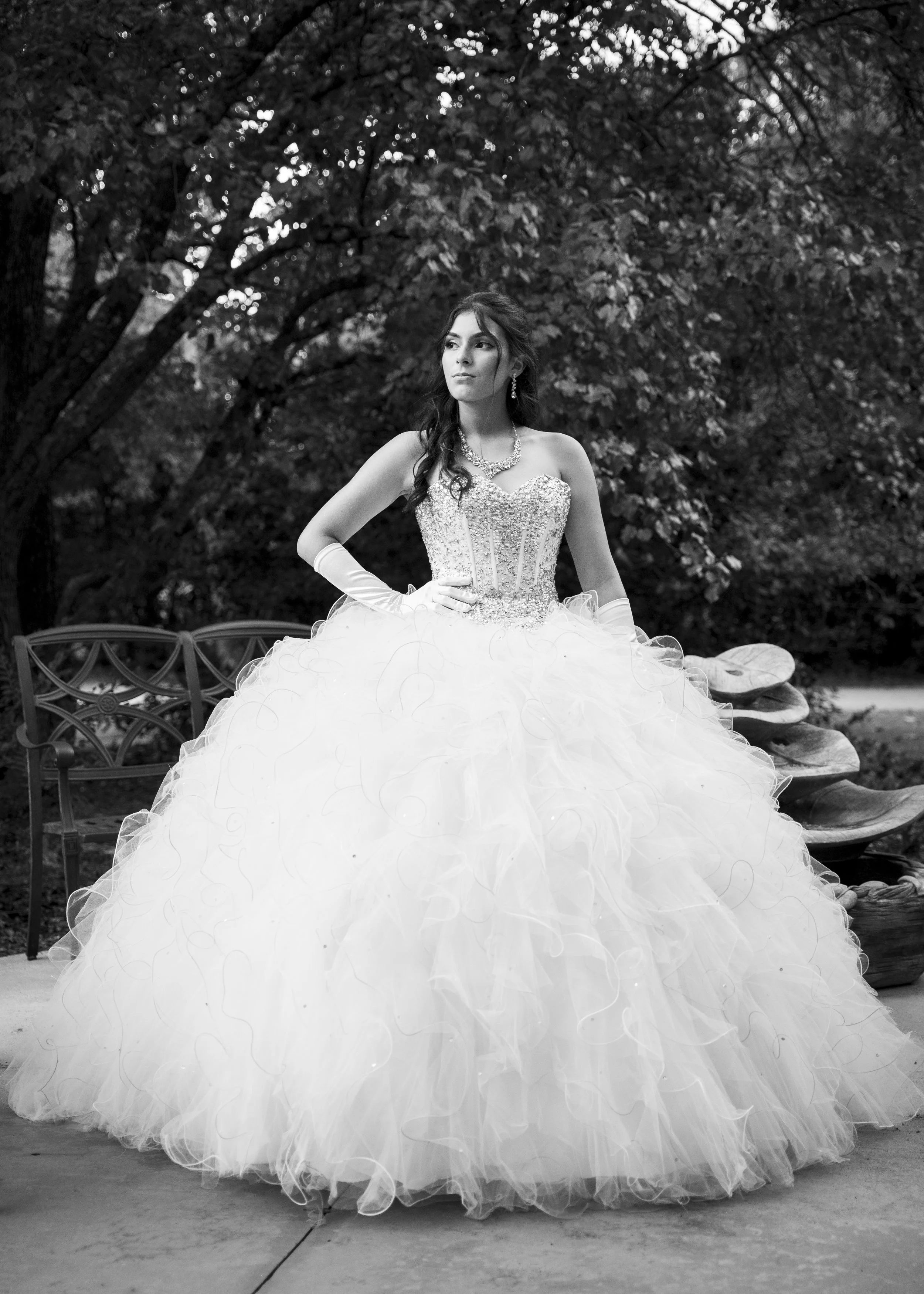 A woman in a strapless, beaded wedding gown with a full, ruffled skirt standing outdoors on a paved area, with trees and a bench in the background.