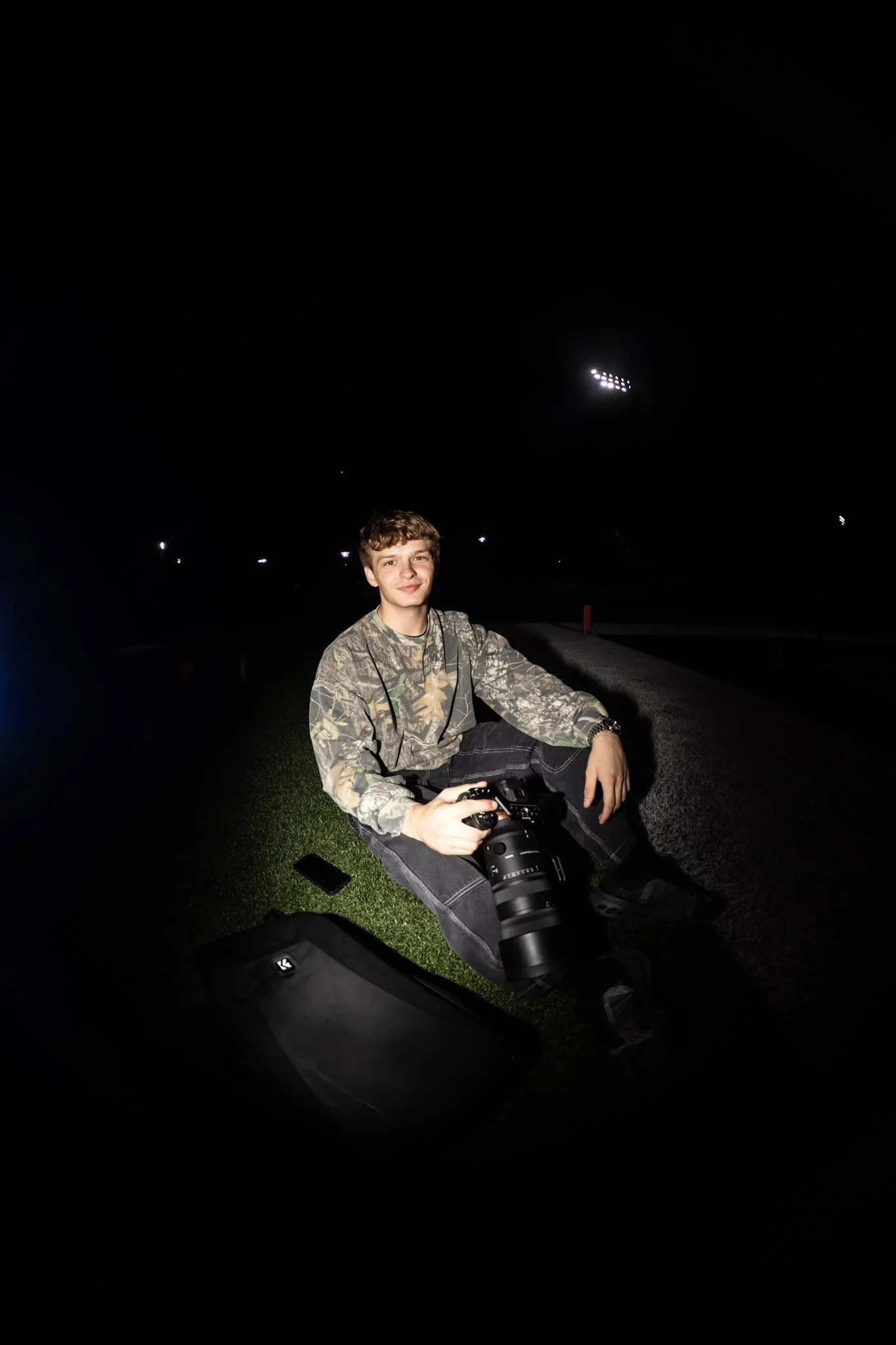 A young man sitting on the grass holding a camera, with a camera bag and mobile phone nearby, outdoors at night.