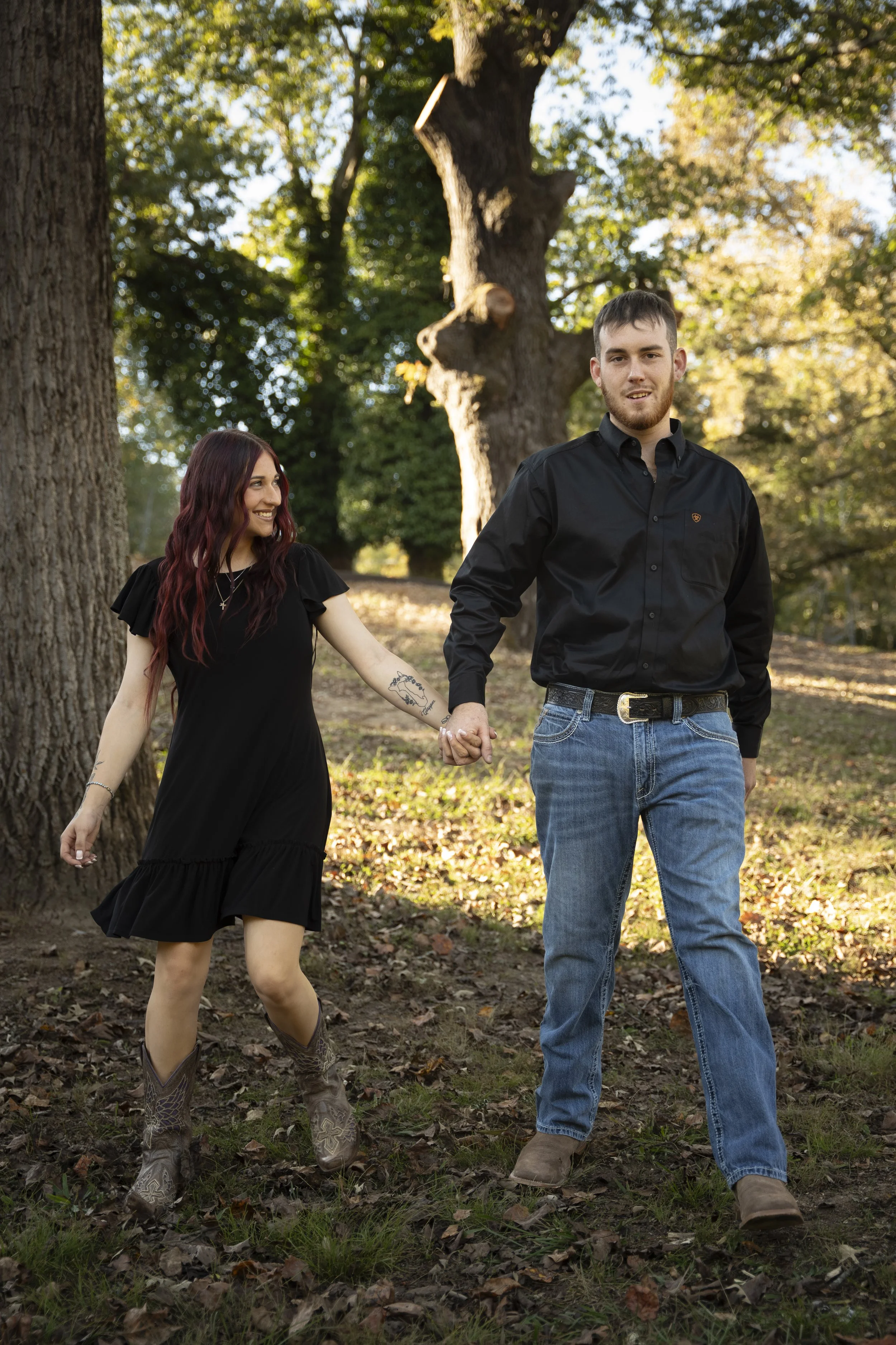 A young couple walking hand-in-hand outdoors in a wooded area with trees and fallen leaves, smiling and enjoying each other's company.