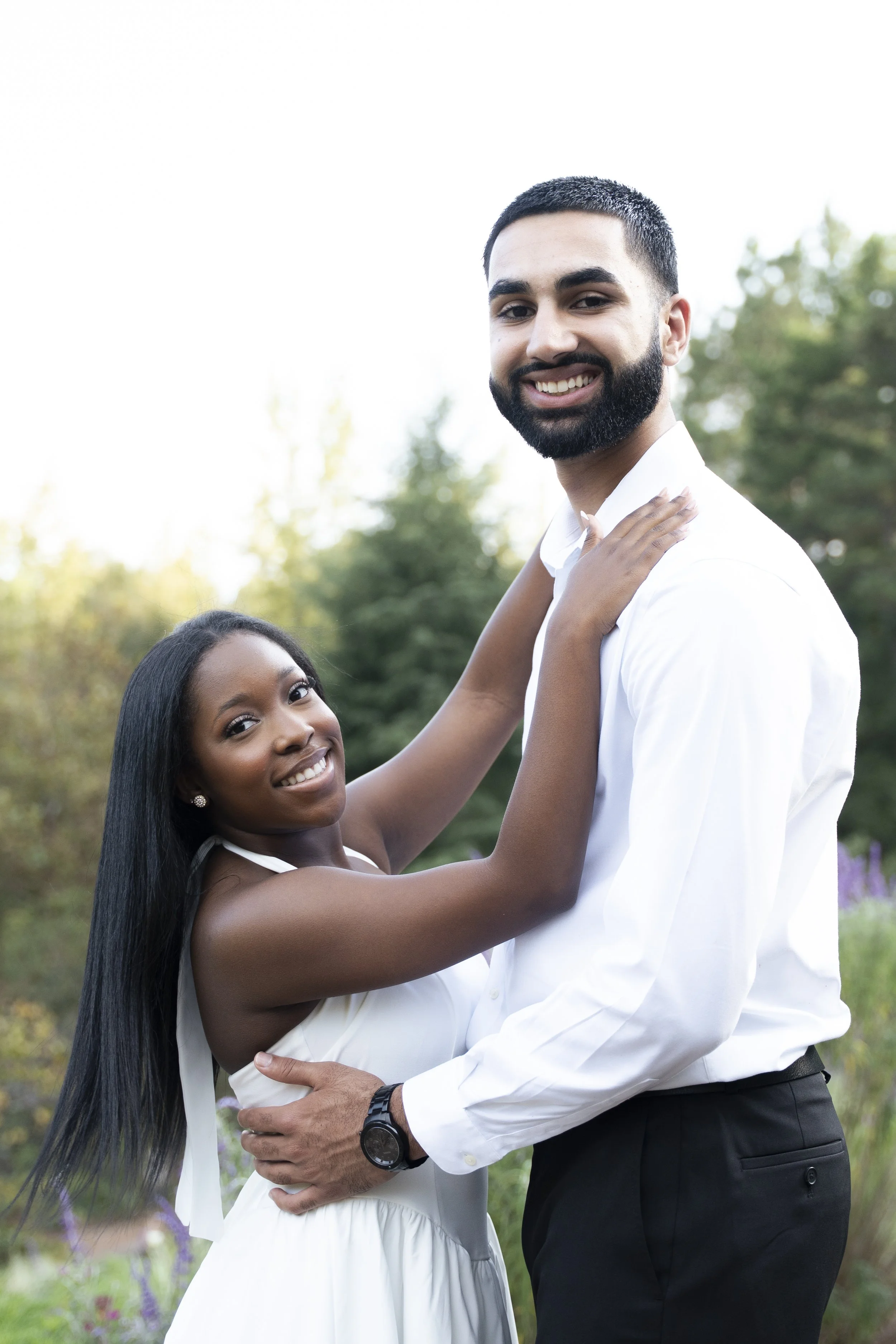 A happy couple standing outdoors, smiling at the camera, with the woman gently touching the man's shoulder and the man's arm around her waist, surrounded by trees and greenery.