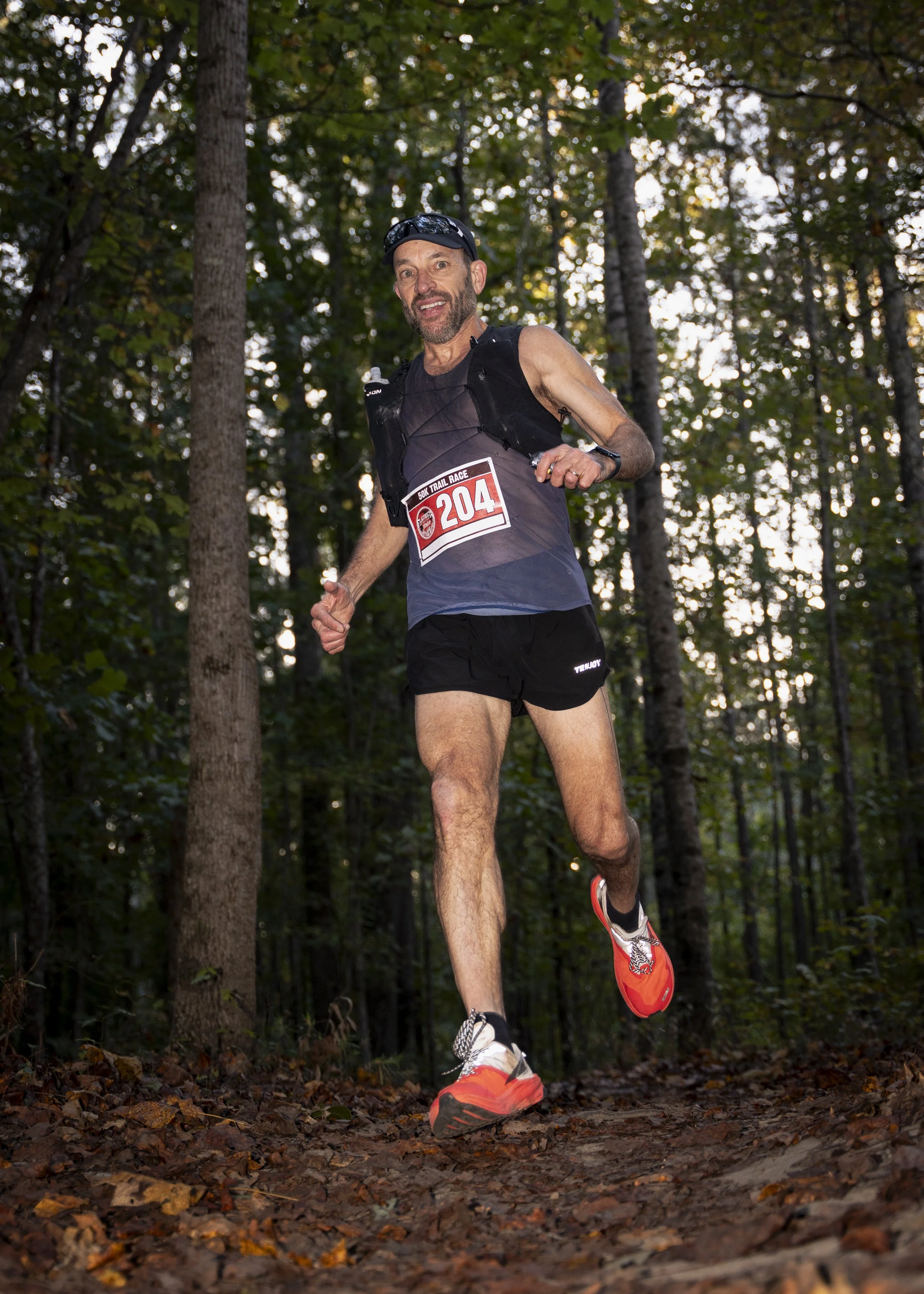 A male trail runner wearing a black tank top, black shorts, orange running shoes, and a race bib numbered 204, running on a leaf-covered forest trail during daytime.