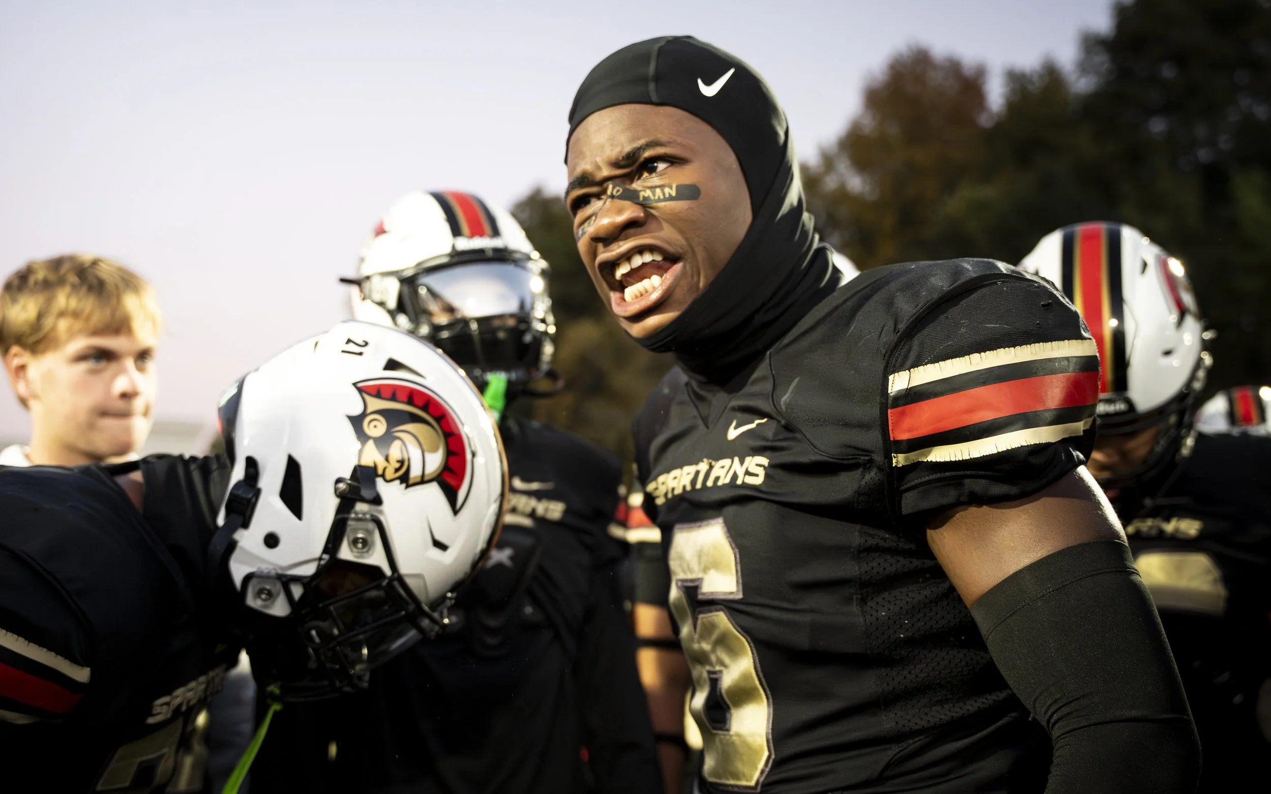 Football player wearing a black jersey with red and gold stripes, black helmet, and face paint in a team huddle on the field.