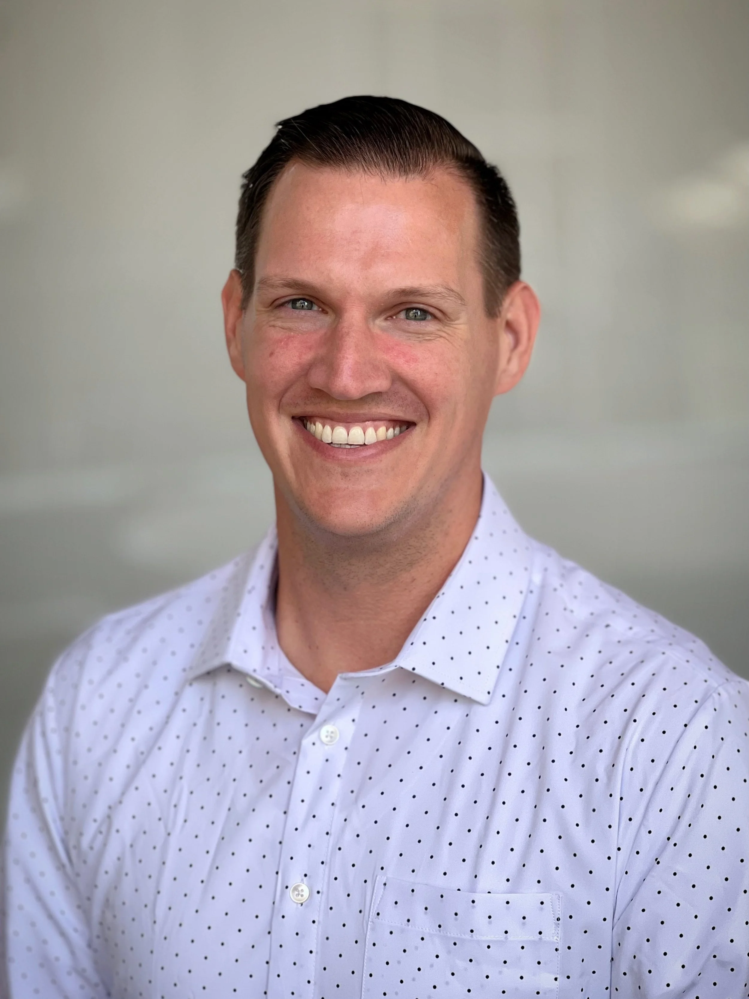 A smiling man with short dark hair wearing a white dress shirt with small black polka dots.