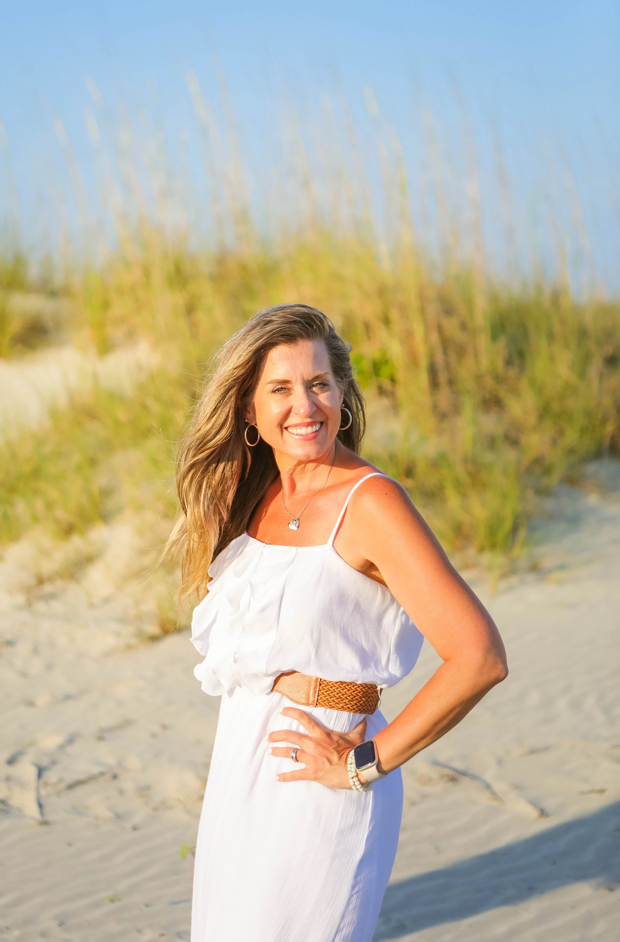 A woman wearing a white dress standing outdoors on a sandy beach with grass in the background. She has long hair, hoop earrings, a necklace, a smartwatch, a ring, and a bracelet, and she is smiling.