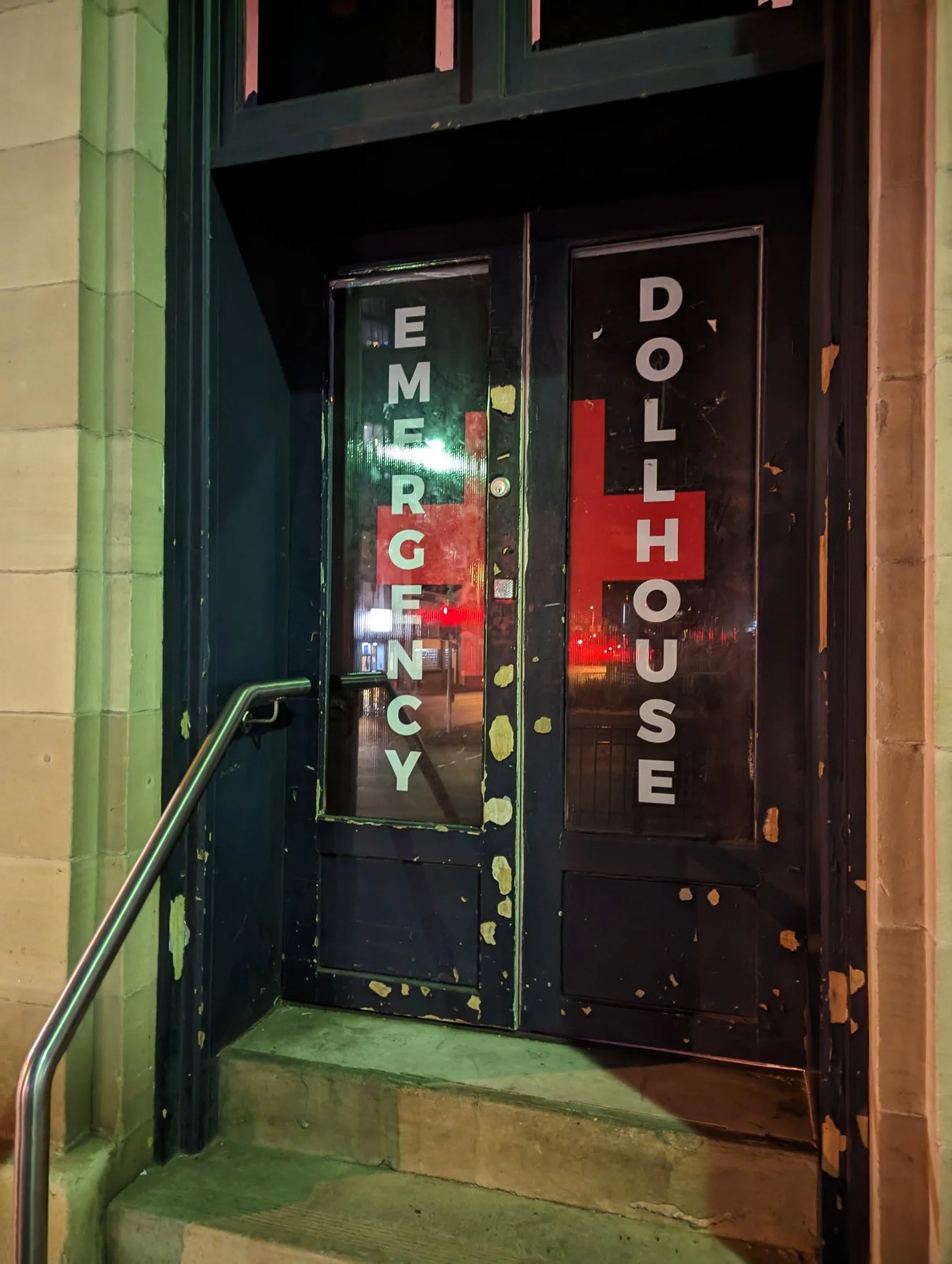 Black door with vertical glass panel featuring white letters spelling "EMERGENCY DOOR" and a partially visible red sign behind the glass. The door is slightly weathered with chipped paint and surrounded by a concrete entrance with a handrail.