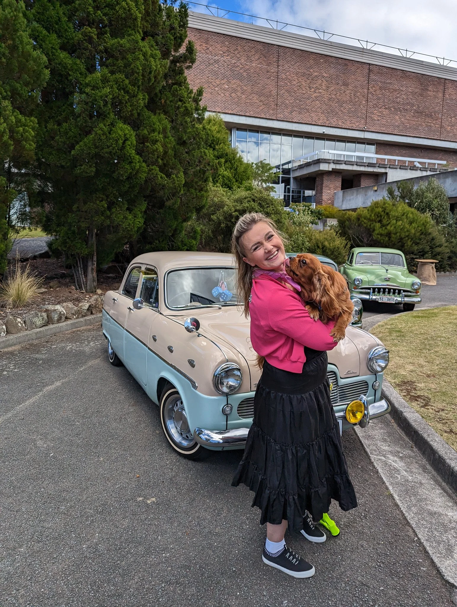 A woman with a big smile holding a brown puppy in front of vintage cars, including a beige classic car with a blue dice hanging from the rearview mirror and a green older model car, parked outside near trees and a large brick building.