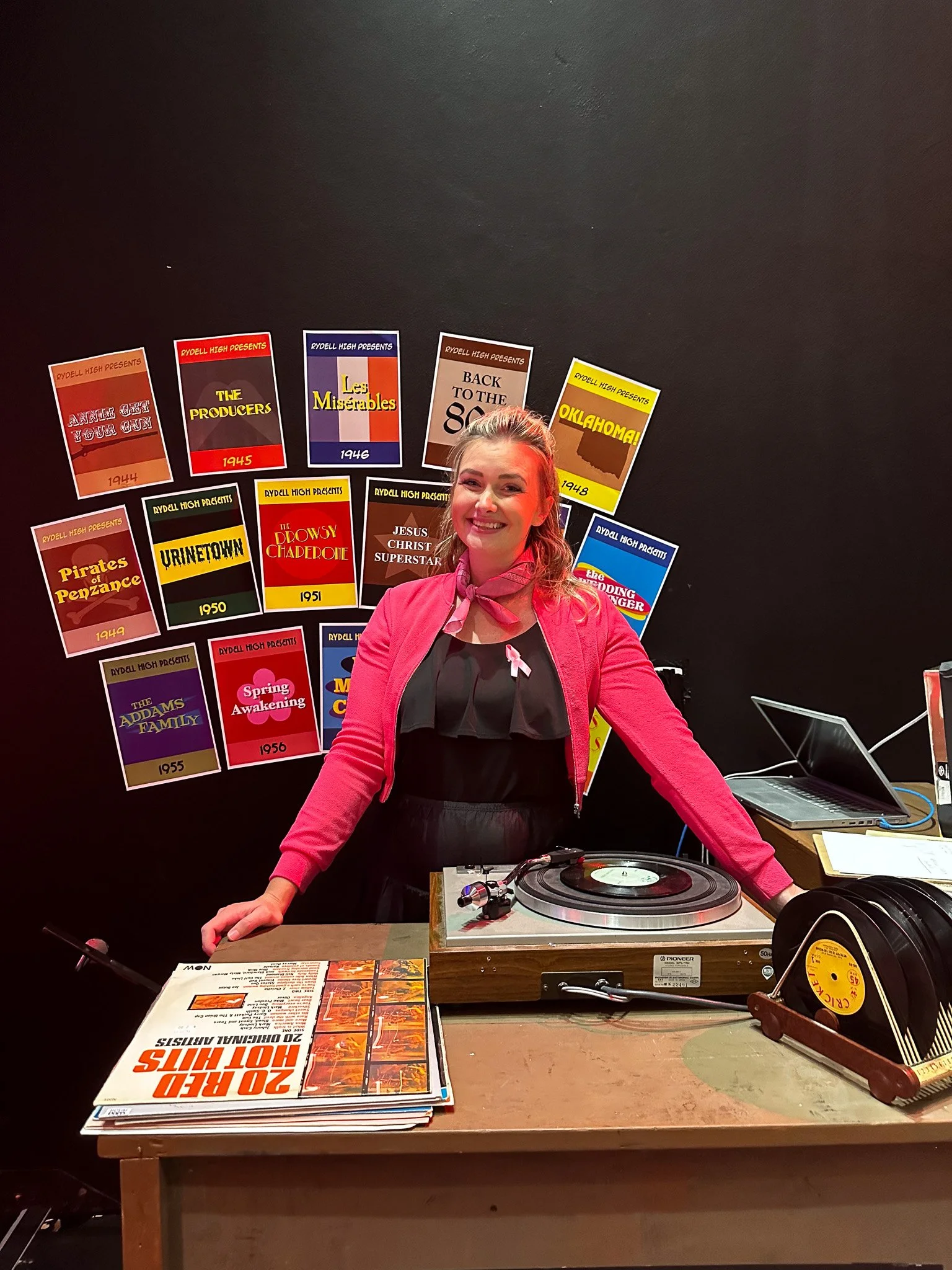 A woman is standing behind a table with a record player, vinyl records, and a menu. Behind her, there are colorful event posters on a black wall.