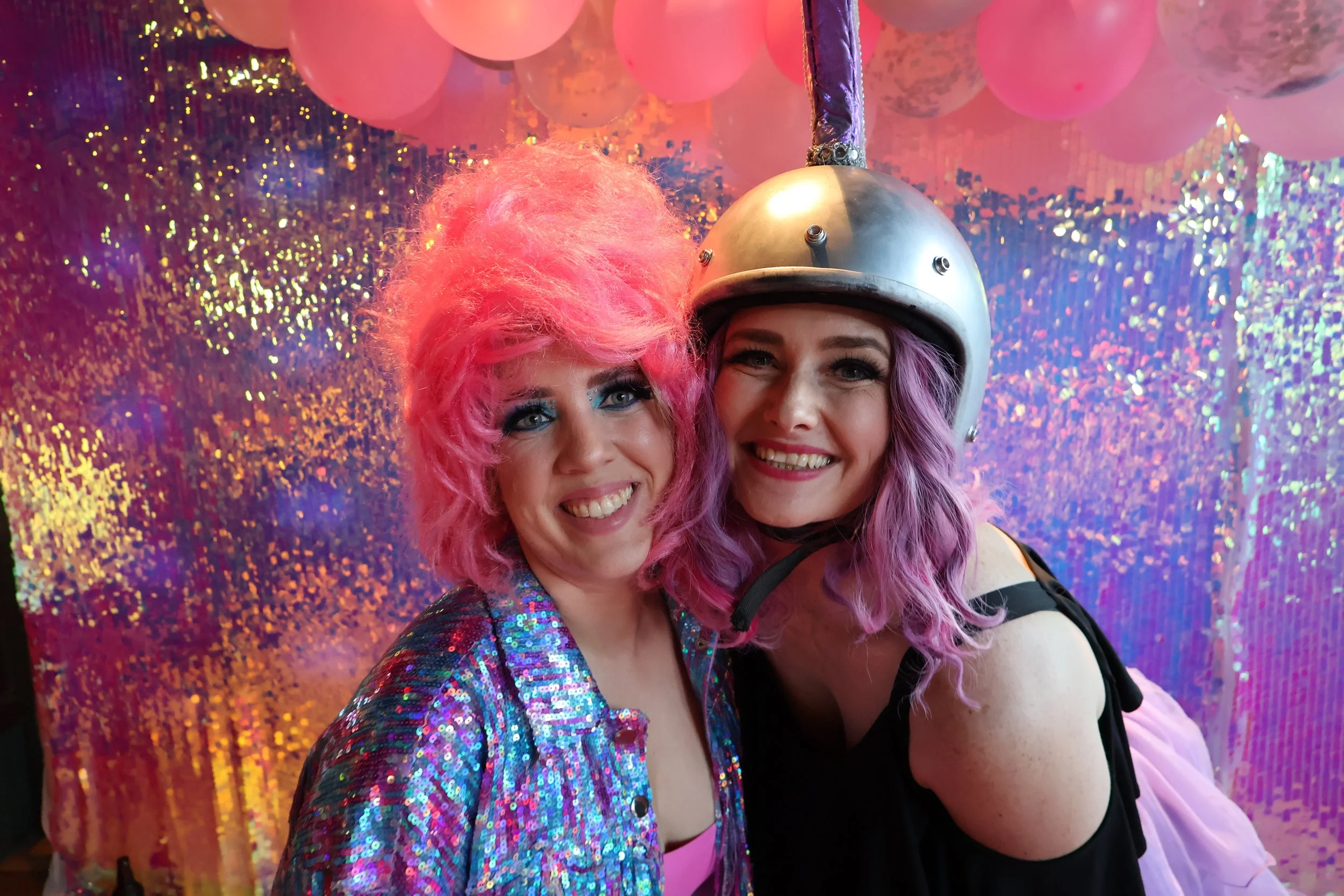 Two women with pink and purple hairstyles taking a selfie at a vibrant, glittery party. One woman is wearing a sequined jacket and has bright pink hair, while the other is wearing a metallic helmet with a unicorn horn and has purple hair. The backgro