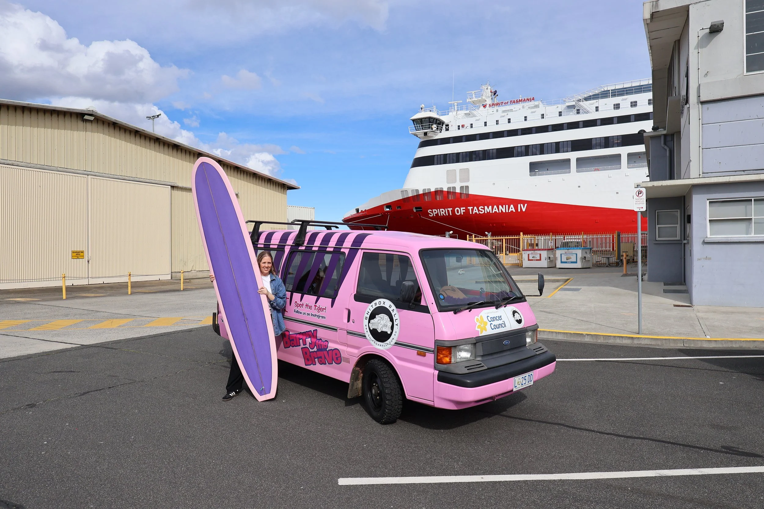 A pink van with the logo 'Barn Run the Brave' and Cancer Council sticker, parked in a lot with a woman holding a purple surfboard, background shows a large ship named 'Spirit of Tasmania IV' and an industrial building.