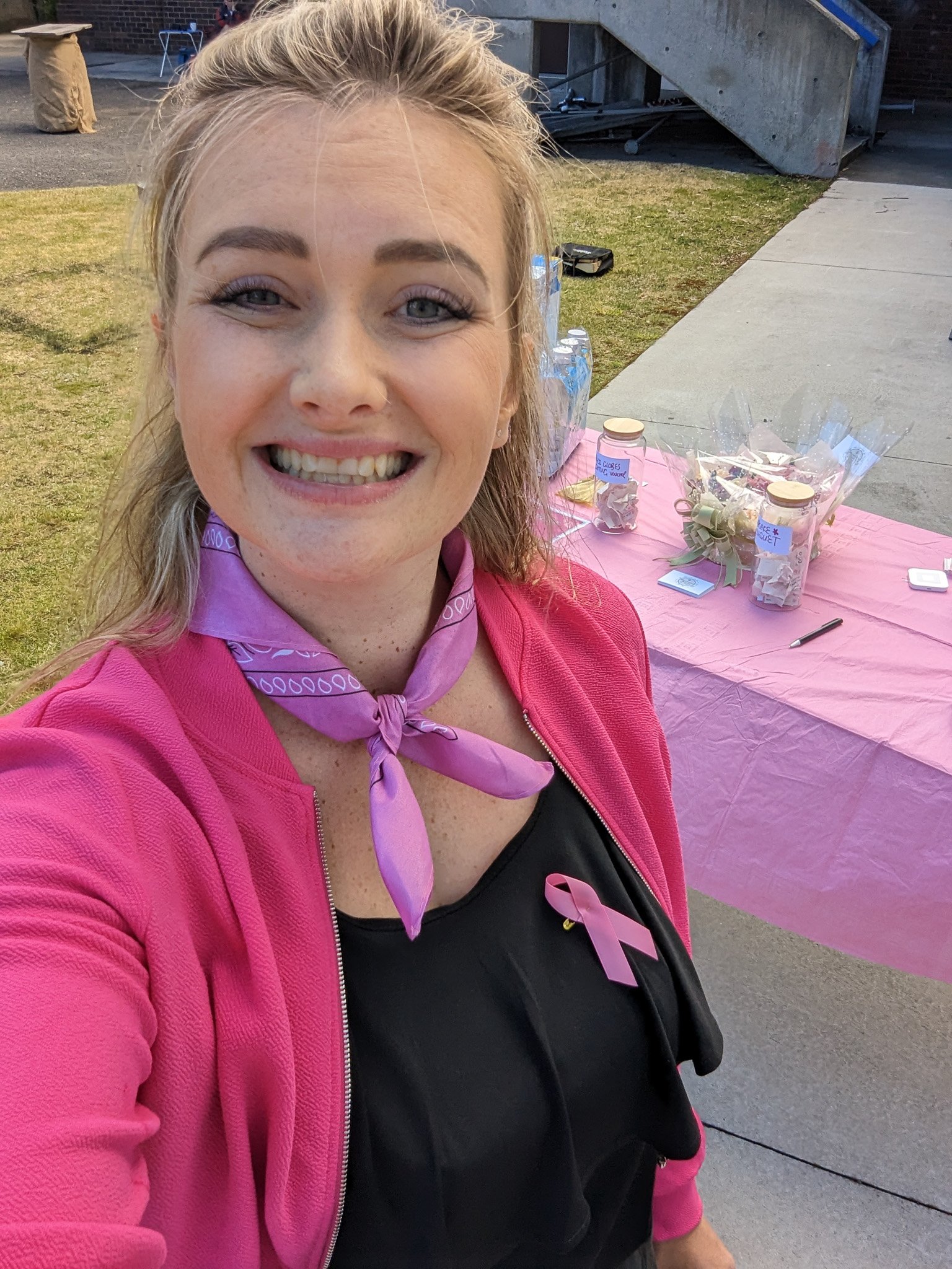 A woman smiling for a selfie, wearing a pink jacket and purple bandana with breast cancer awareness ribbon, at an outdoor event with tables and water bottles in background.