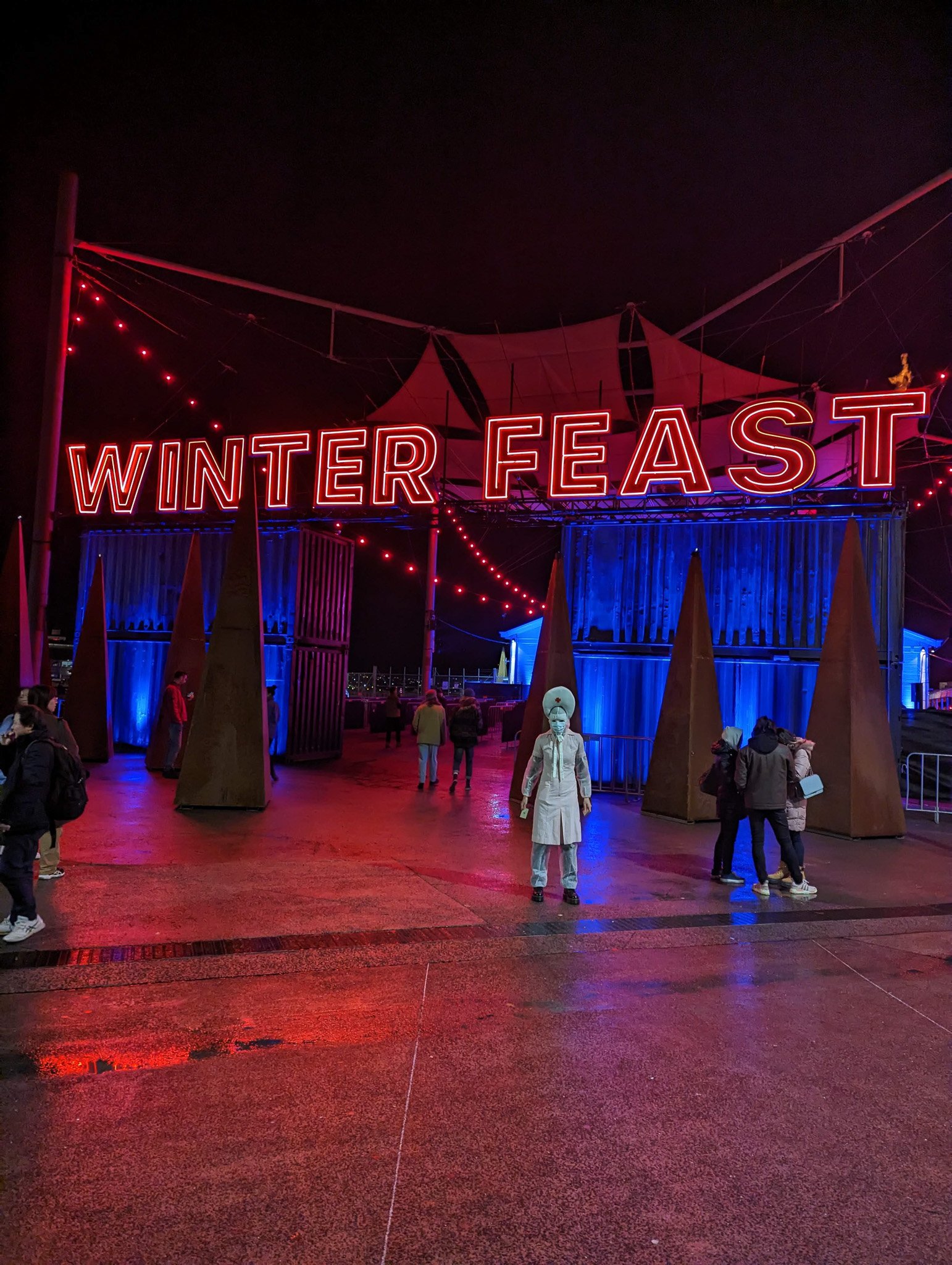 Night scene at Winter Feast with a large illuminated sign, people walking, and decorative blue lighting on structures, illuminated by red and blue lights.