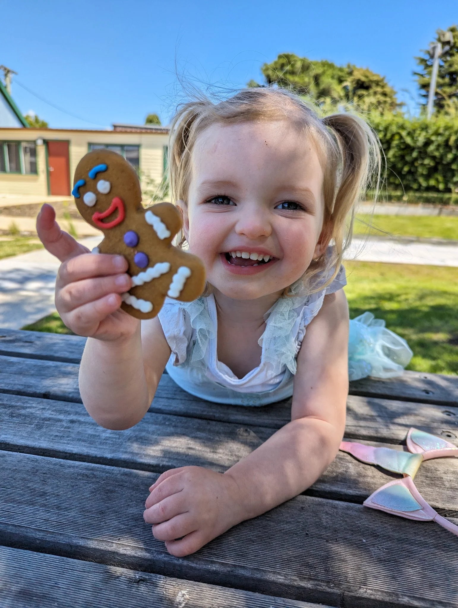 A young girl with blonde hair in pigtails holding a gingerbread cookie with colorful icing, smiling outdoors on a wooden surface with a house, green bushes, and blue sky in the background.