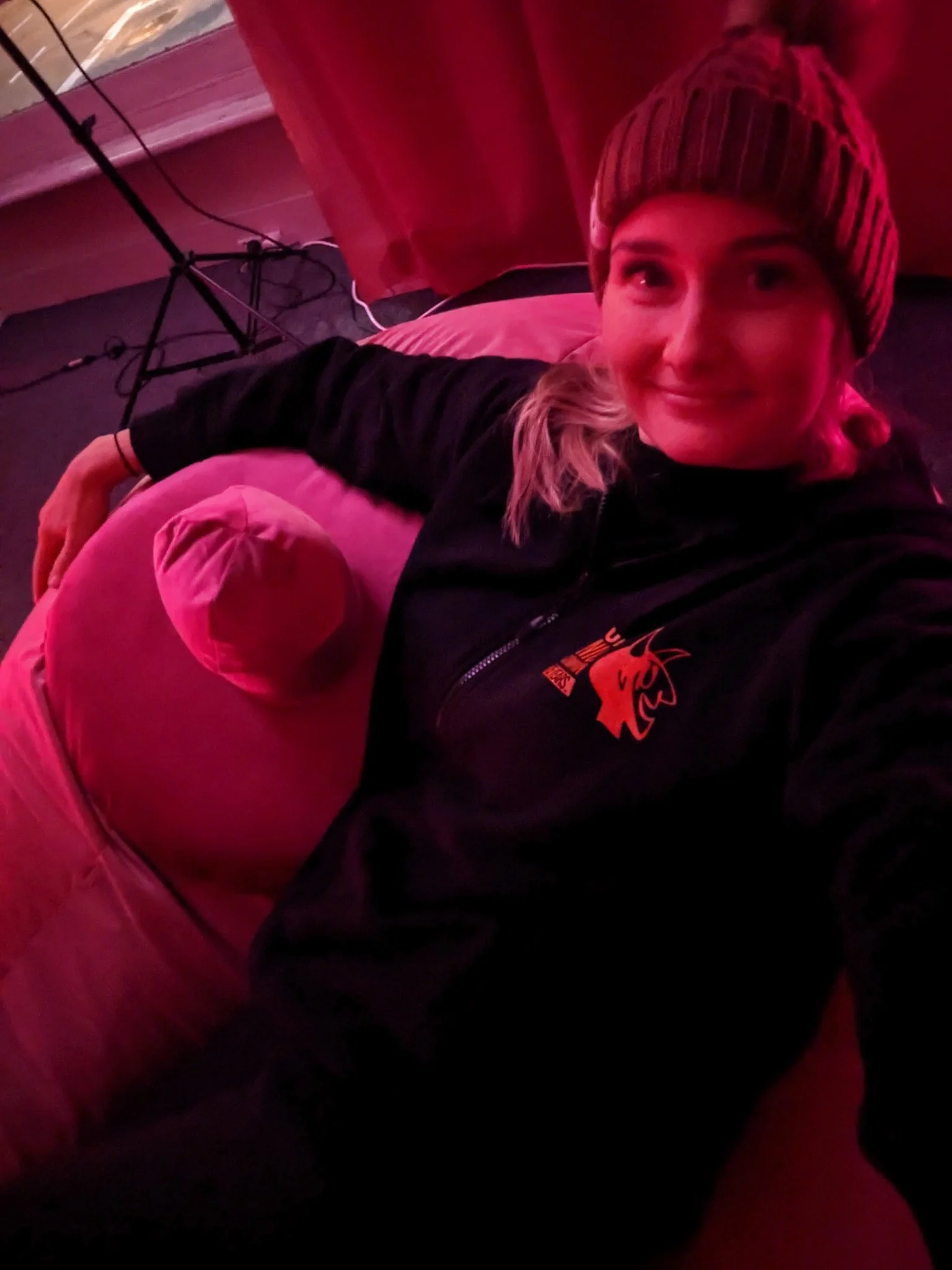 A person lying in bed with a smile, wearing a striped beanie and a jacket while taking a selfie in a room with a red curtain and a black floor lamp.