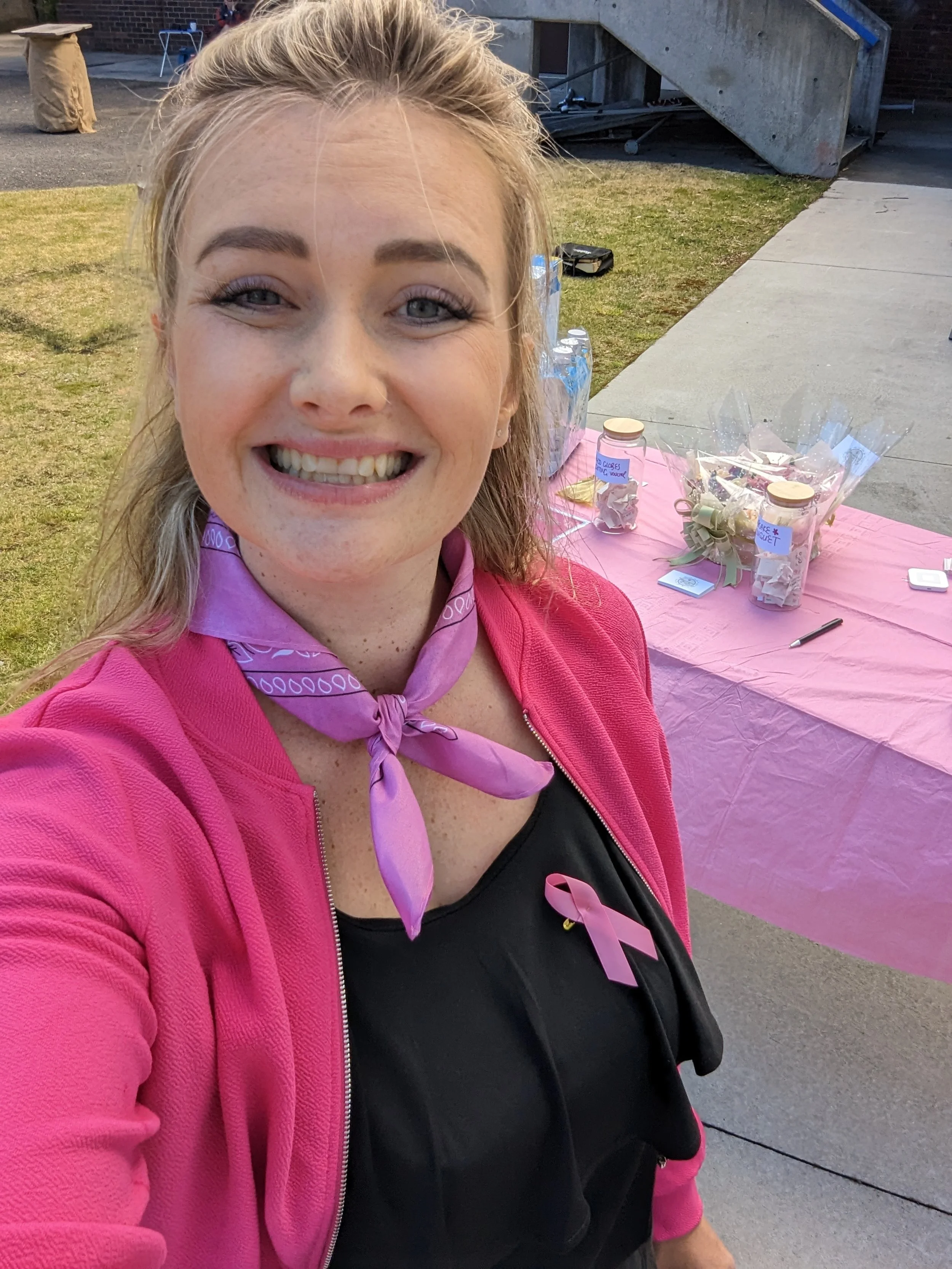 A smiling woman with blonde hair and gray eyes wearing a pink jacket, a purple neckerchief, and a black shirt with a pink ribbon, taking a selfie outdoors in front of a table with small gift bags, jars, and water bottles.