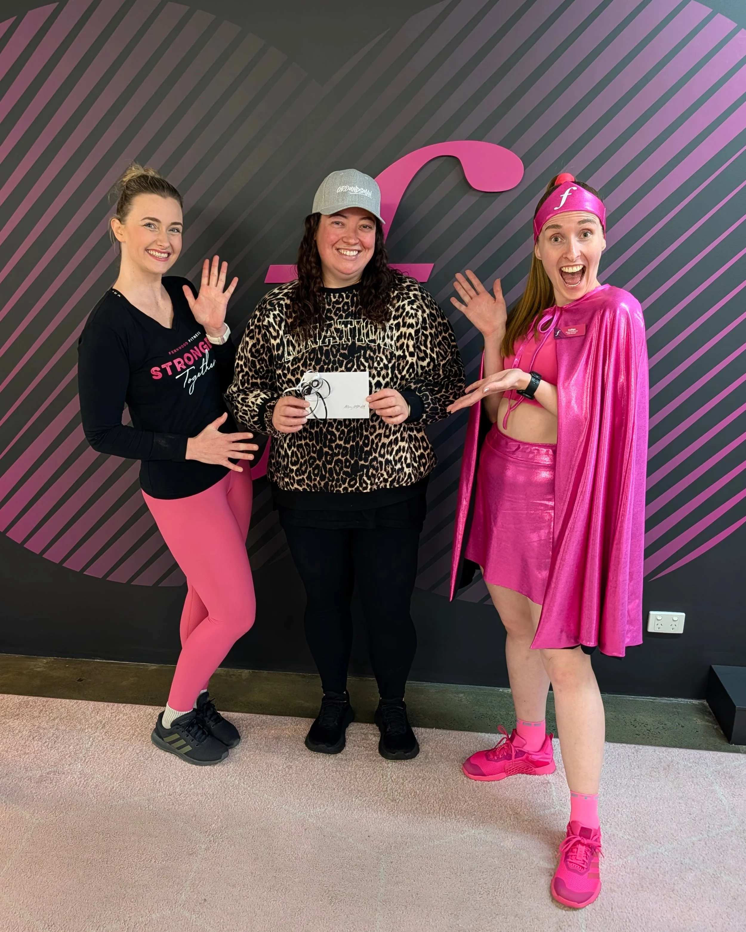 Three women posing together indoors in front of a dark wall with pink stripes and a large pink logo. The woman on the left is wearing a black long-sleeve shirt and pink leggings, smiling and waving. The woman in the middle is wearing a leopard print 
