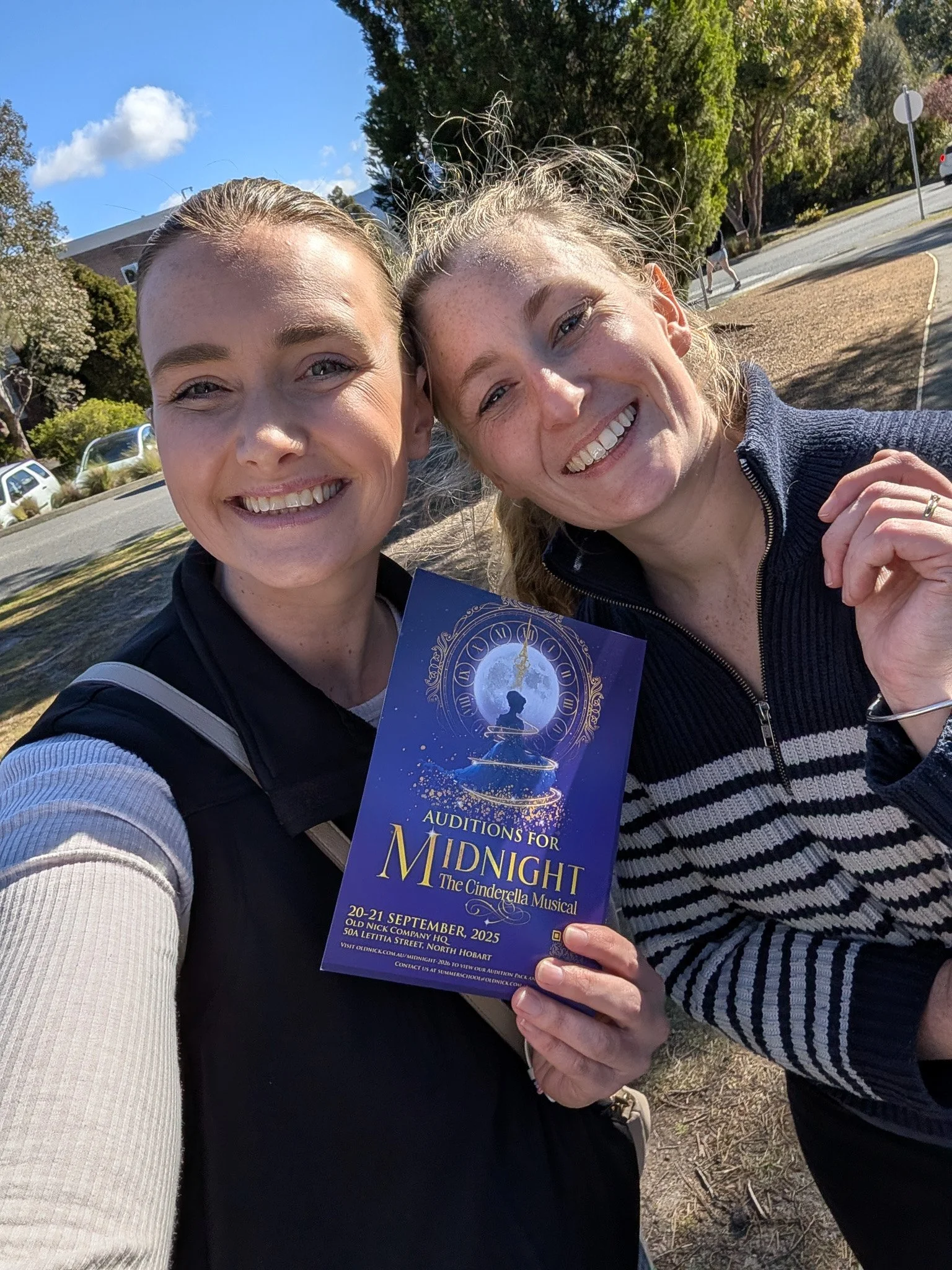 Two women smiling, taking a selfie outdoors in sunny weather, holding a flyer for auditions for 'Midnight: The Cinderella Musical' happening September 20-21, 2025, in North Hobart, Australia.