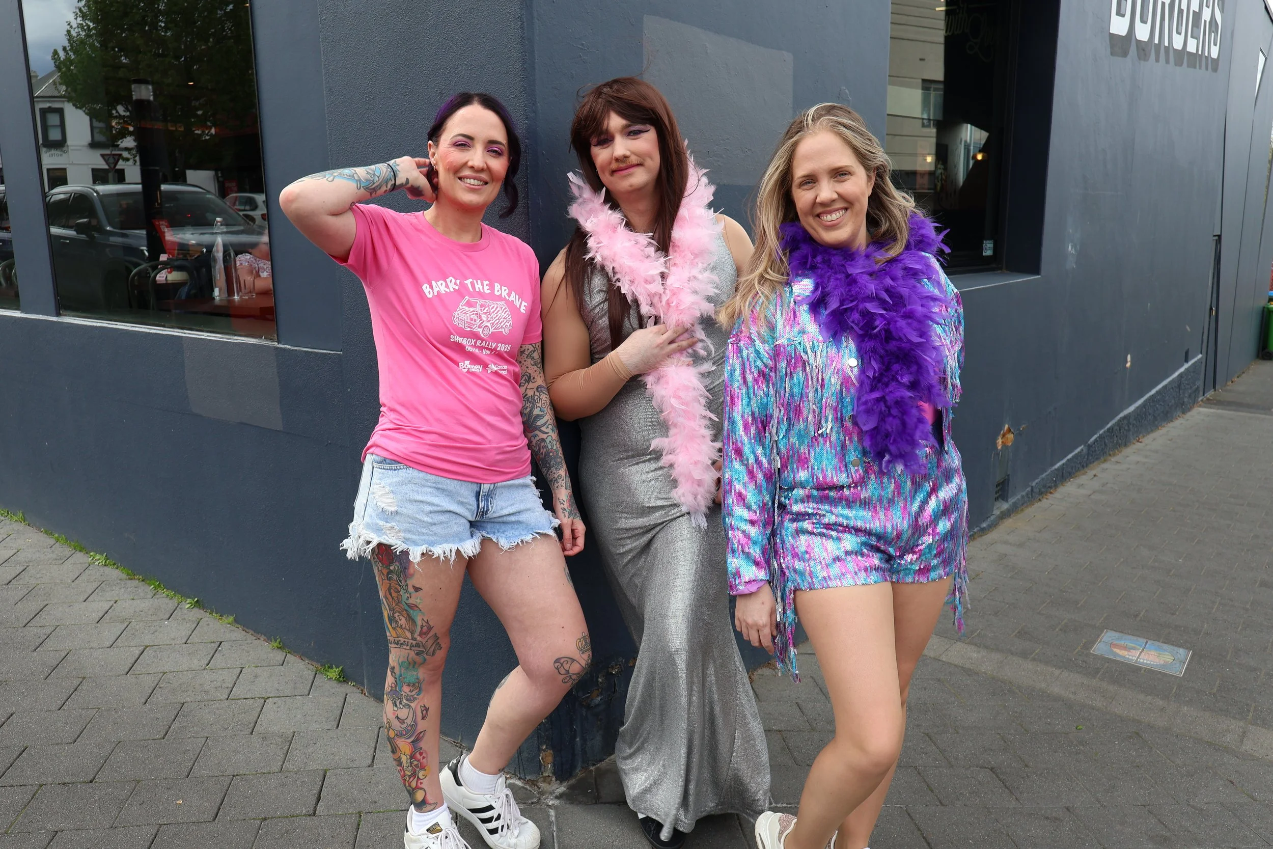 Three women standing outside in front of a dark gray wall, smiling and posing for a photo. The woman on the left has dark hair, tattoos, and is wearing a pink t-shirt and denim shorts. The woman in the middle has brown hair, a feather boa, and a shin