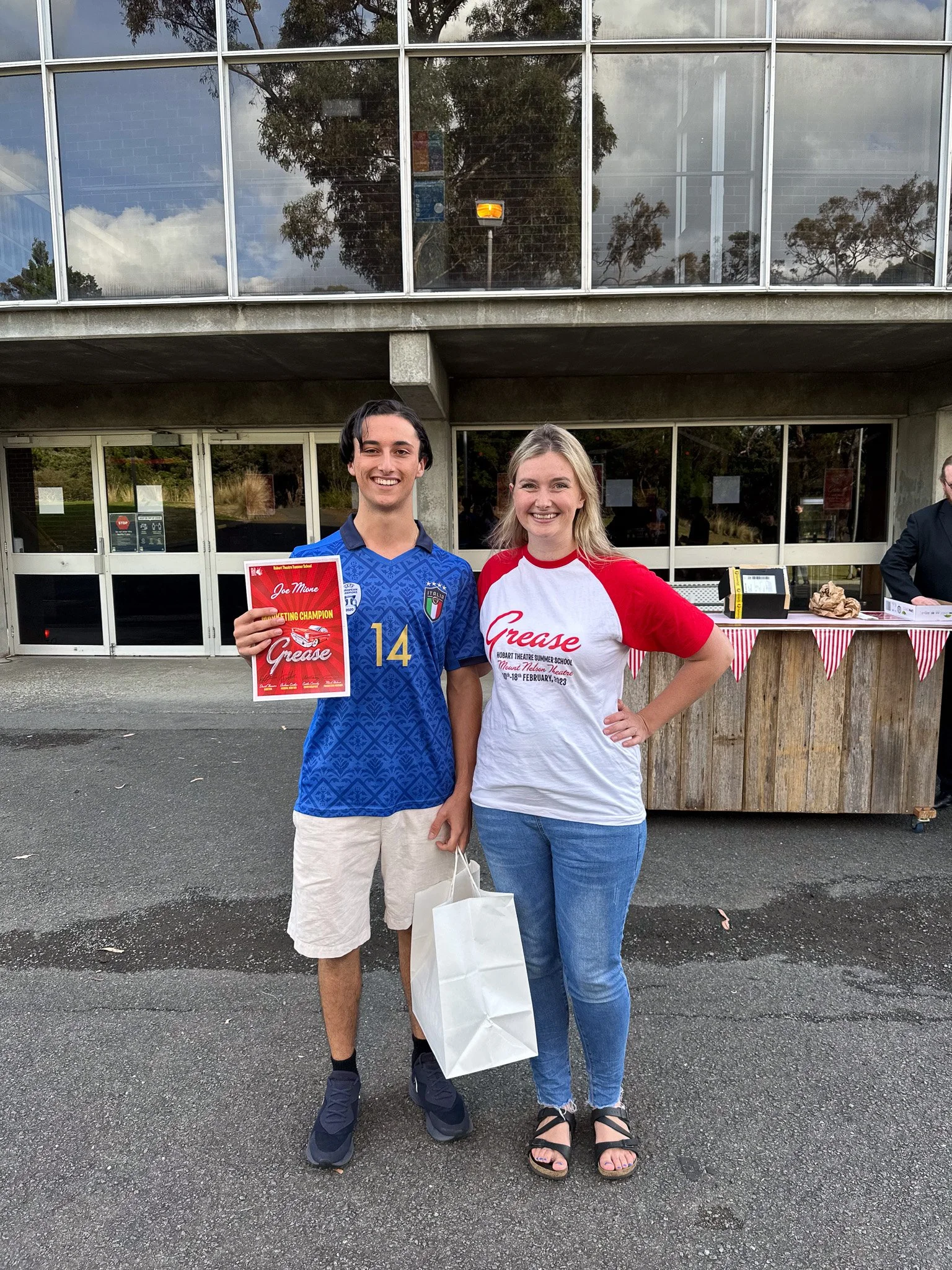 Two young adults, a man and a woman, standing outdoors in front of a building with large glass windows. The man is wearing a blue sports jersey with the Italian flag and holding a red award certificate. The woman is wearing a white and red t-shirt th