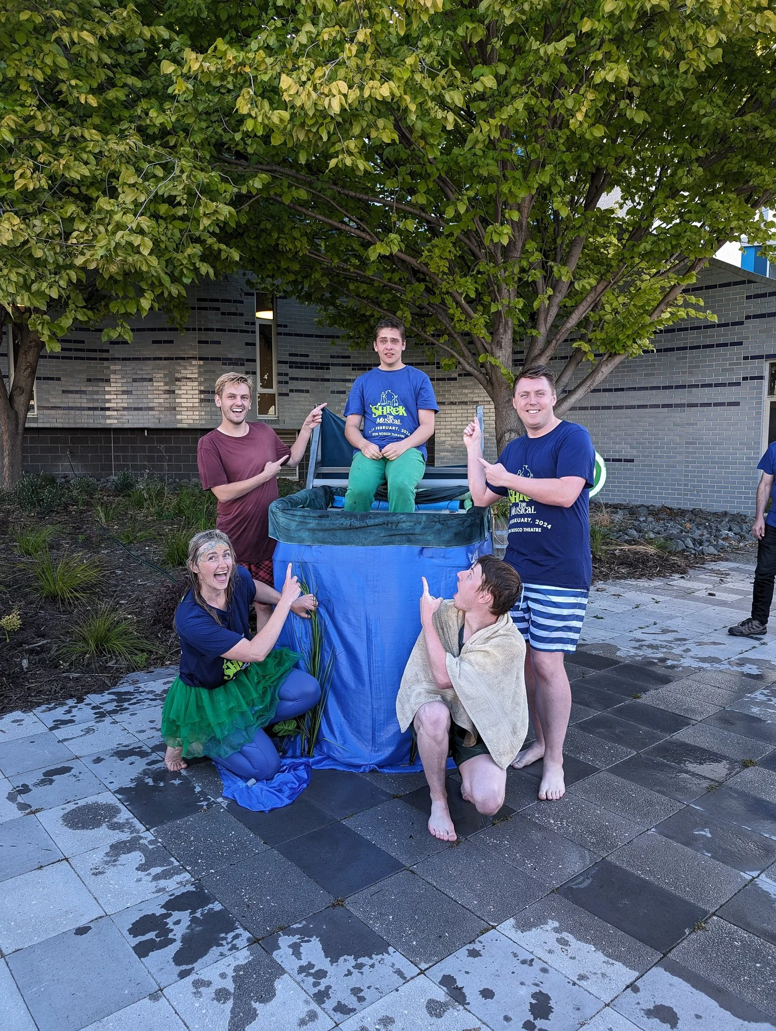 Group of young people outdoors, some in colorful costumes, with a tree and a building in the background. They are smiling and pointing at a makeshift pool or washing container.