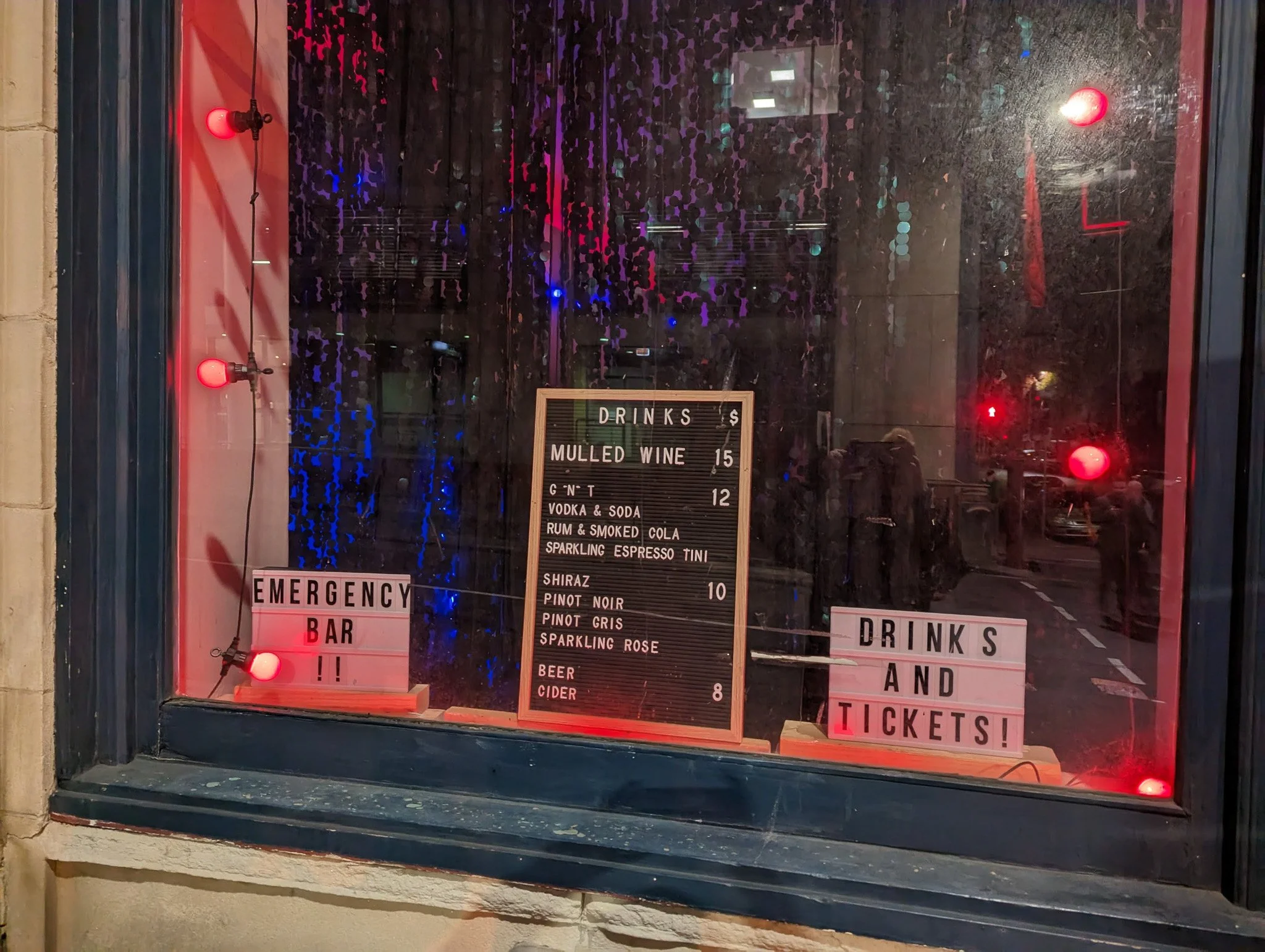 A window display with a black and red color theme, featuring red lights, a black signboard listing drink options such as mulled wine, gin, vodka, rum, and sparkling wine, and various signs indicating an emergency bar and ticketing policies, with refl