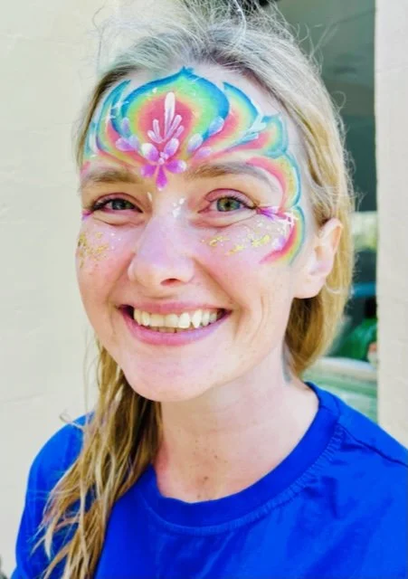 Woman with rainbow and unicorn face paint, smiling, wearing a blue shirt, outdoors near a building