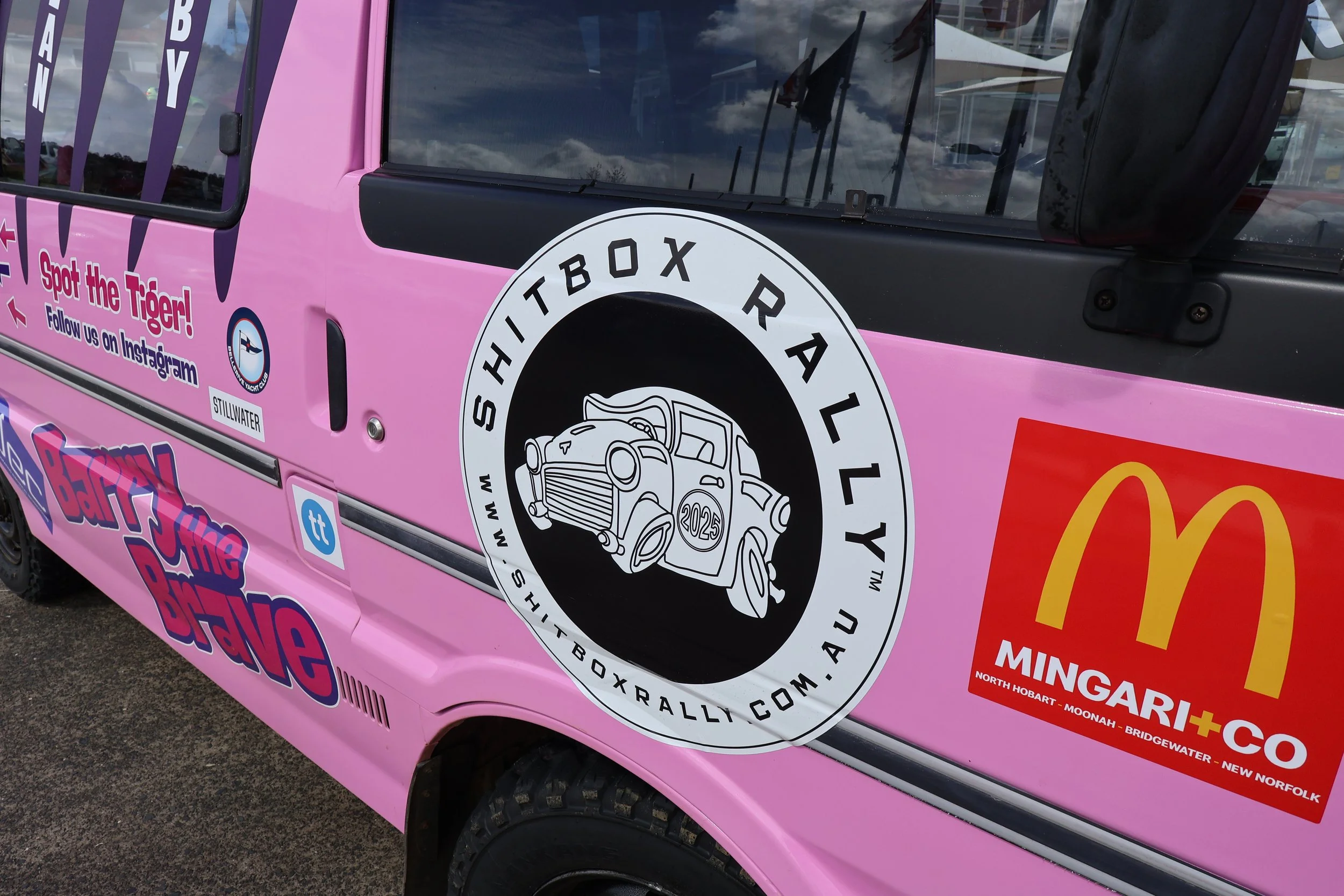 Side of a pink vehicle with various stickers and logos, including a large circular logo for the Sydney Trophy Rally featuring a vintage car illustration, and advertisements for McDonald's and a social media campaign.