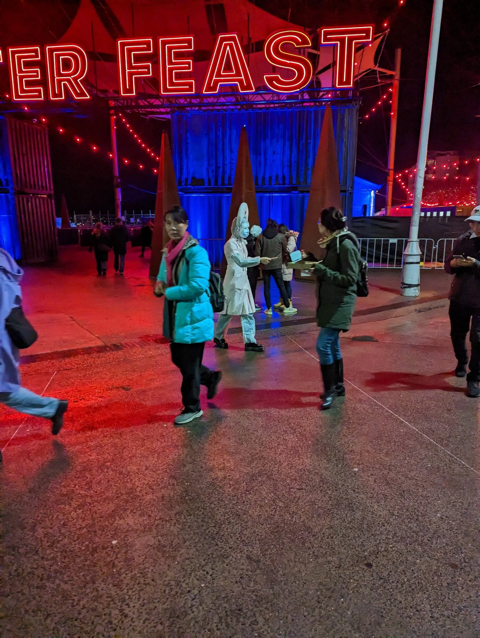 Nighttime scene of a busy city street with people walking. There is a large illuminated sign that reads 'EER FEAST' in red neon lights. The street is wet, reflecting the colorful lights, and there are various people wearing jackets and face masks.