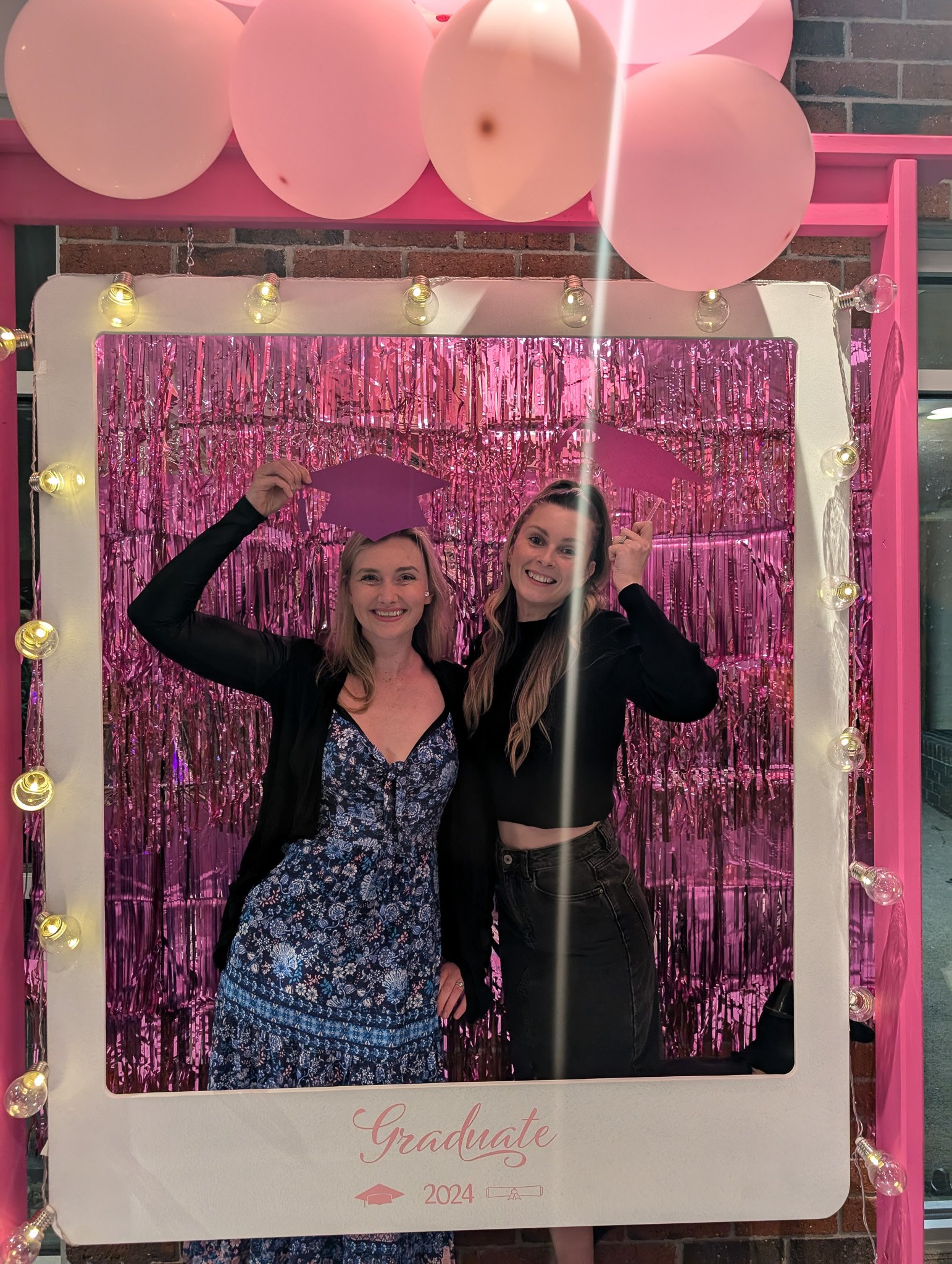 Two women are celebrating graduation, smiling and posing in front of a pink tinsel backdrop. They are holding a graduation cap and a diploma, and are surrounded by pink balloons and graduation-themed decorations.