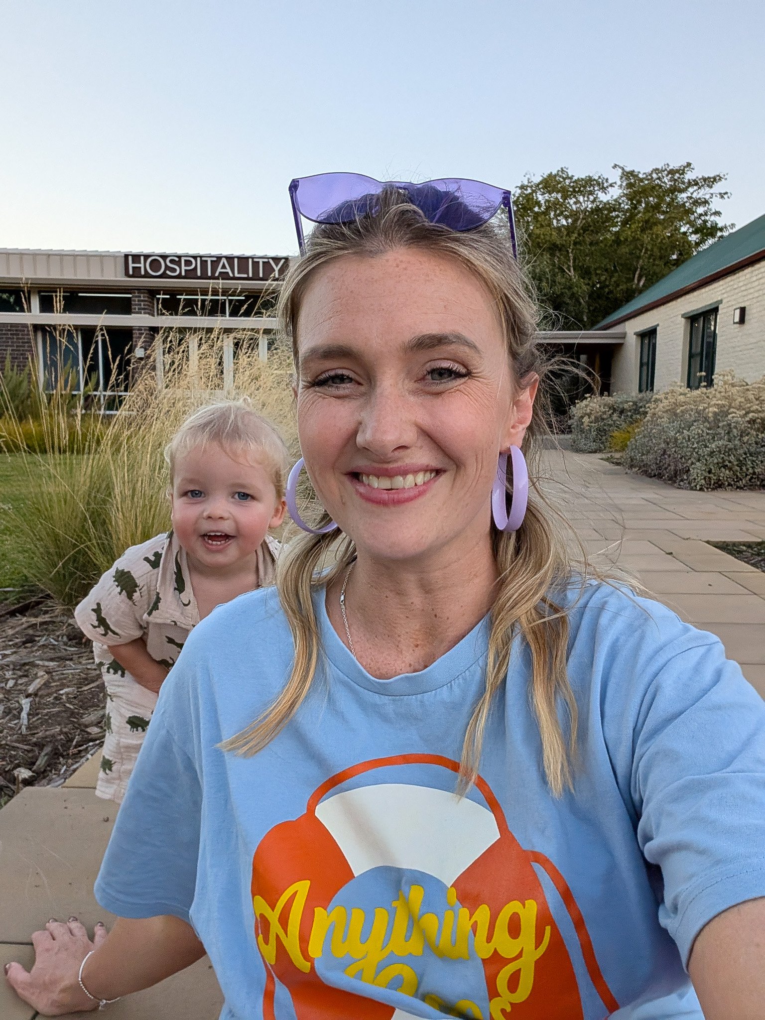 A woman taking a selfie outdoors with a young child behind her, both smiling. The woman is wearing purple sunglasses on her head and a blue T-shirt with colorful text. The background includes a building with a sign that reads 'HOSPITALITY' and some g