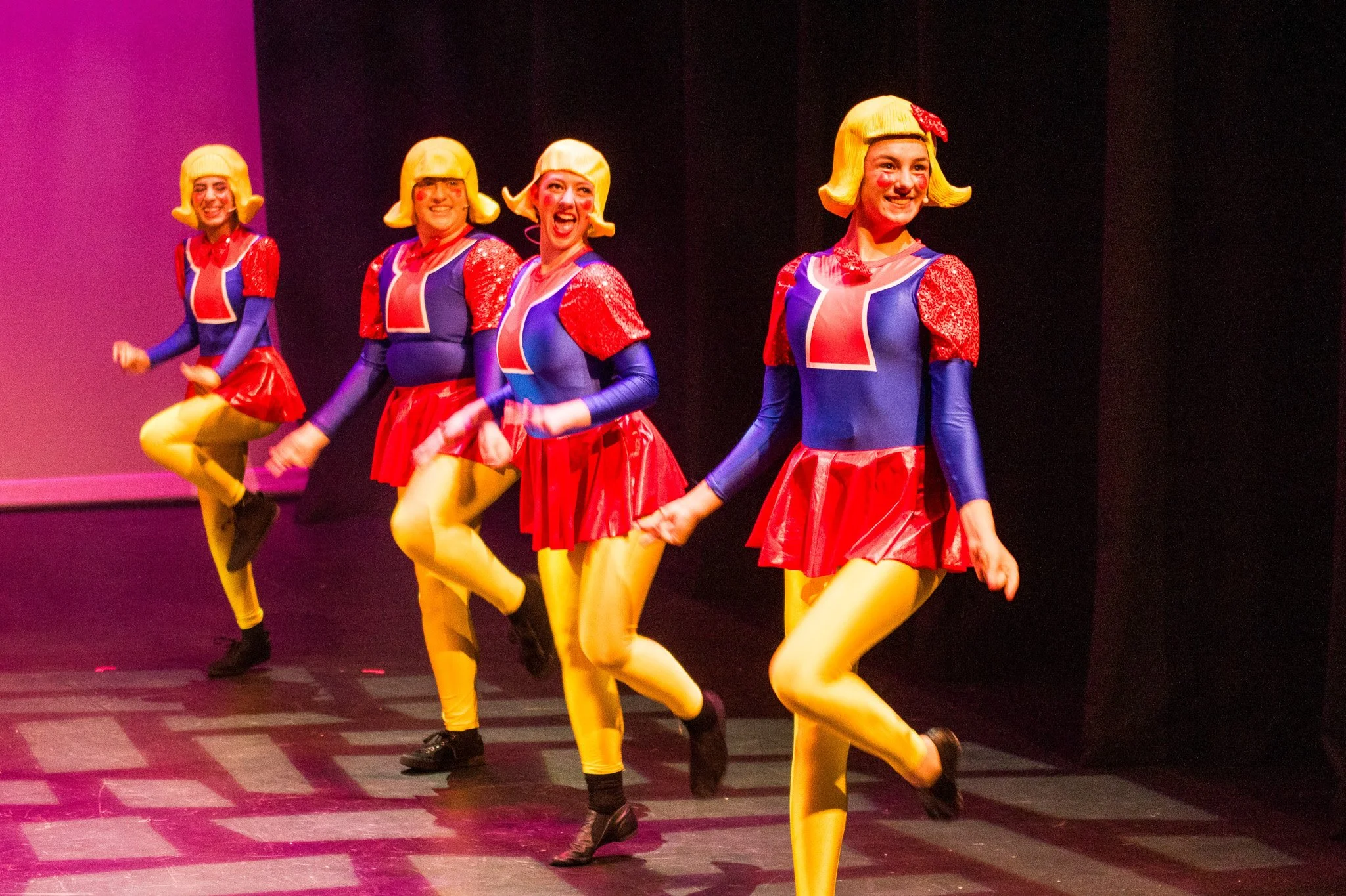Four women dressed in colorful costumes are performing a dance on stage. They are wearing yellow tights, blue tops with red and white accents, red skirts, and yellow wigs with a red bow. They are smiling and appear to be enjoying their performance.