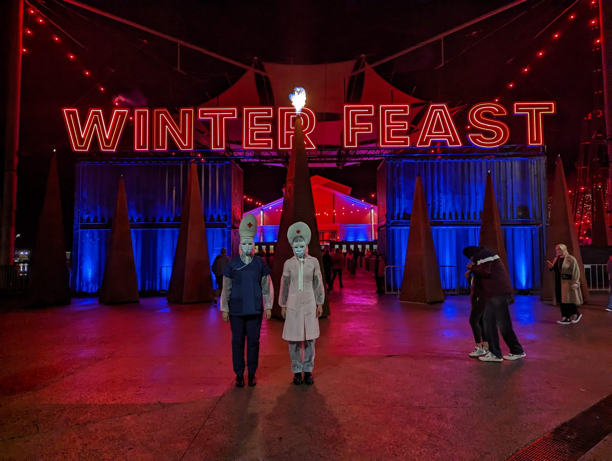 Night scene at an event called Winter Feast with a large neon sign displaying the event's name, people wearing face masks, some in nurse costumes, and illuminated tents with red and blue lighting.