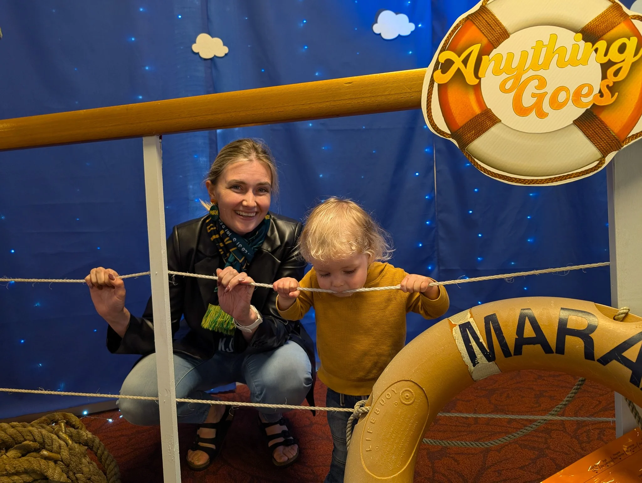 A woman and two children posing behind a pirate-themed photo frame at a party or event. The frame has the words 'Anything Goes' with clouds and blue background decorations.