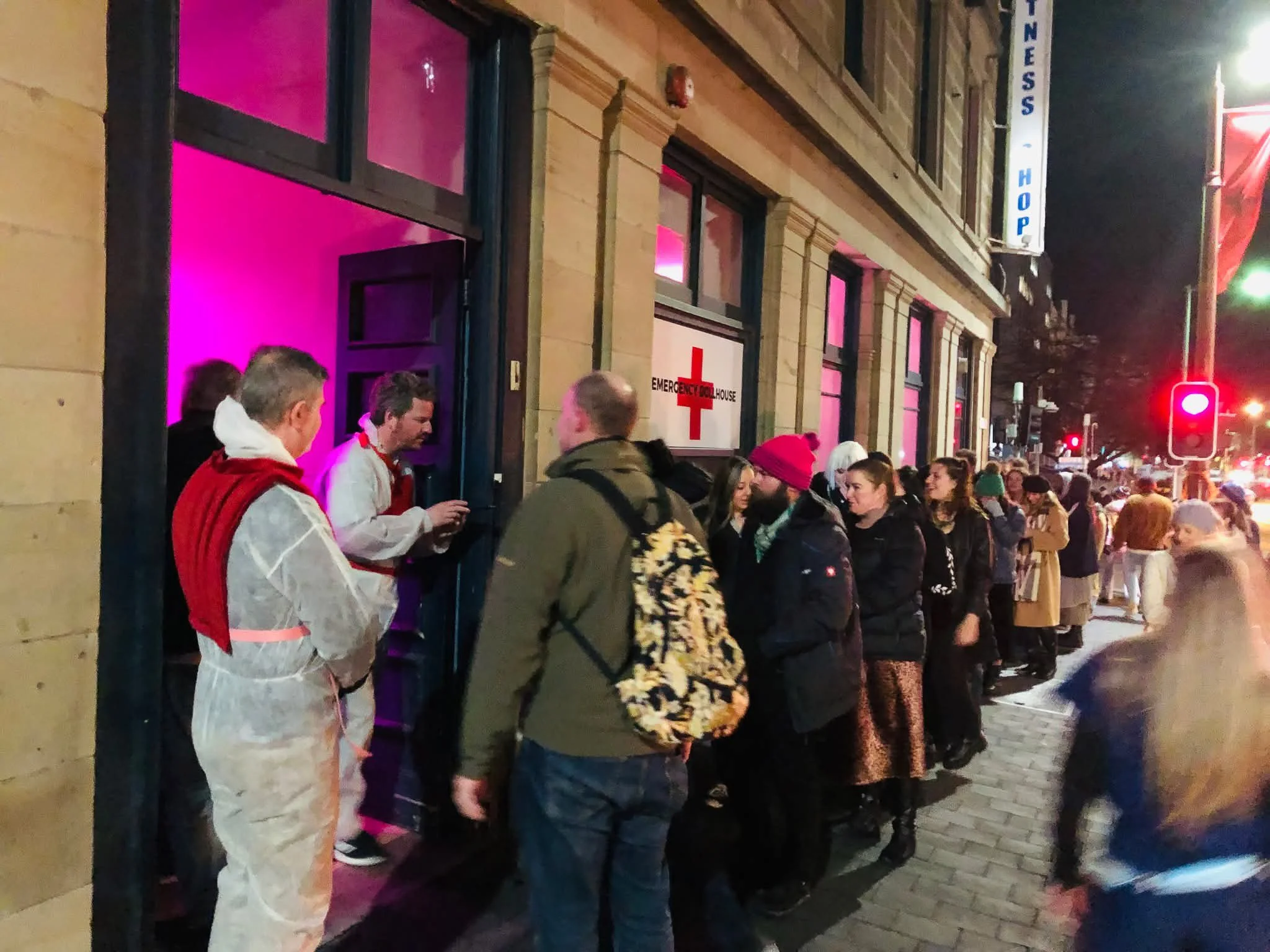 People standing in line outside a building with pink lighting, near an emergency house sign, on a city street at night.