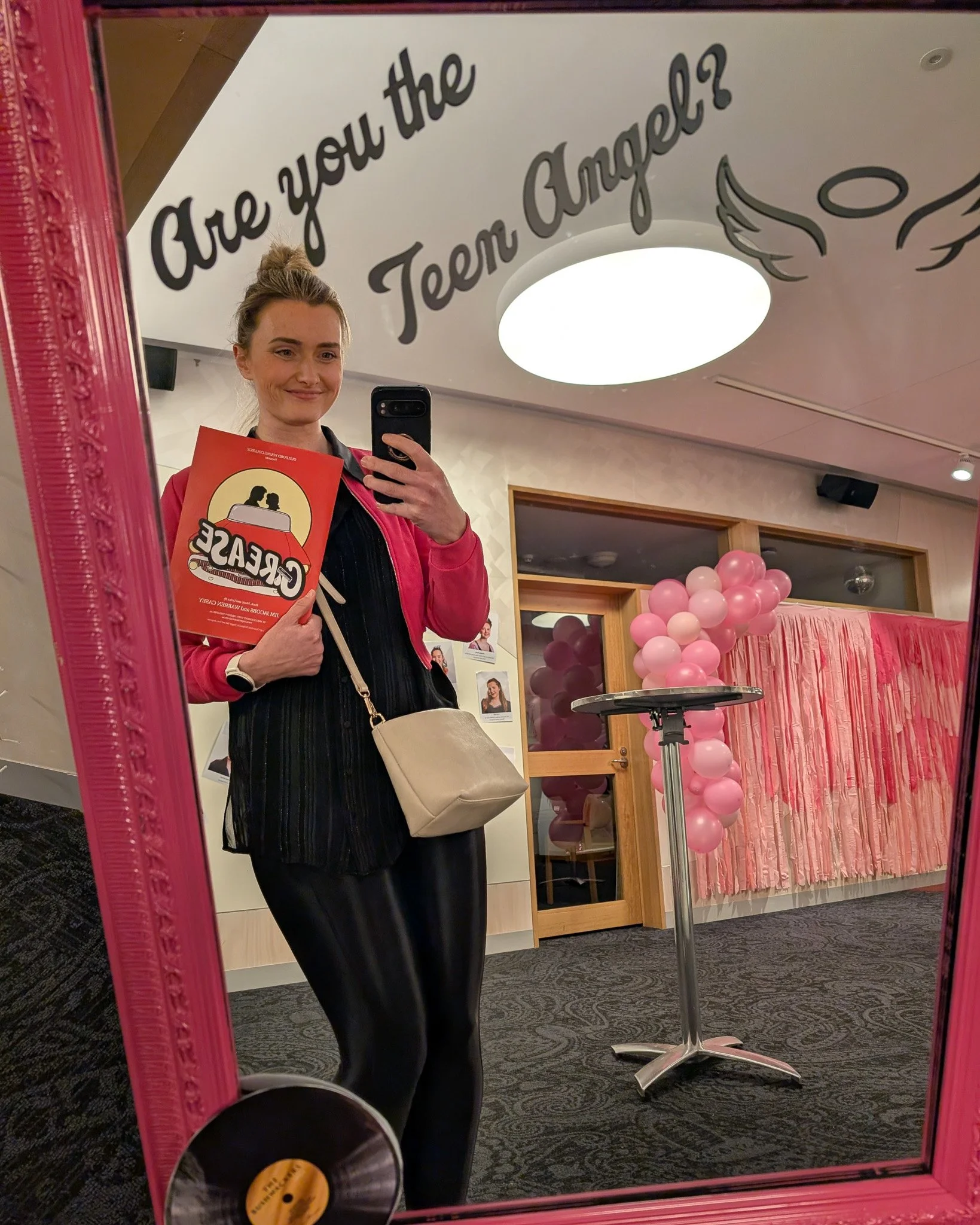 A woman taking a mirror selfie at a celebration event, holding a red party flyer, with pink balloons and a pink curtain in the background, and a wall with photos of children.