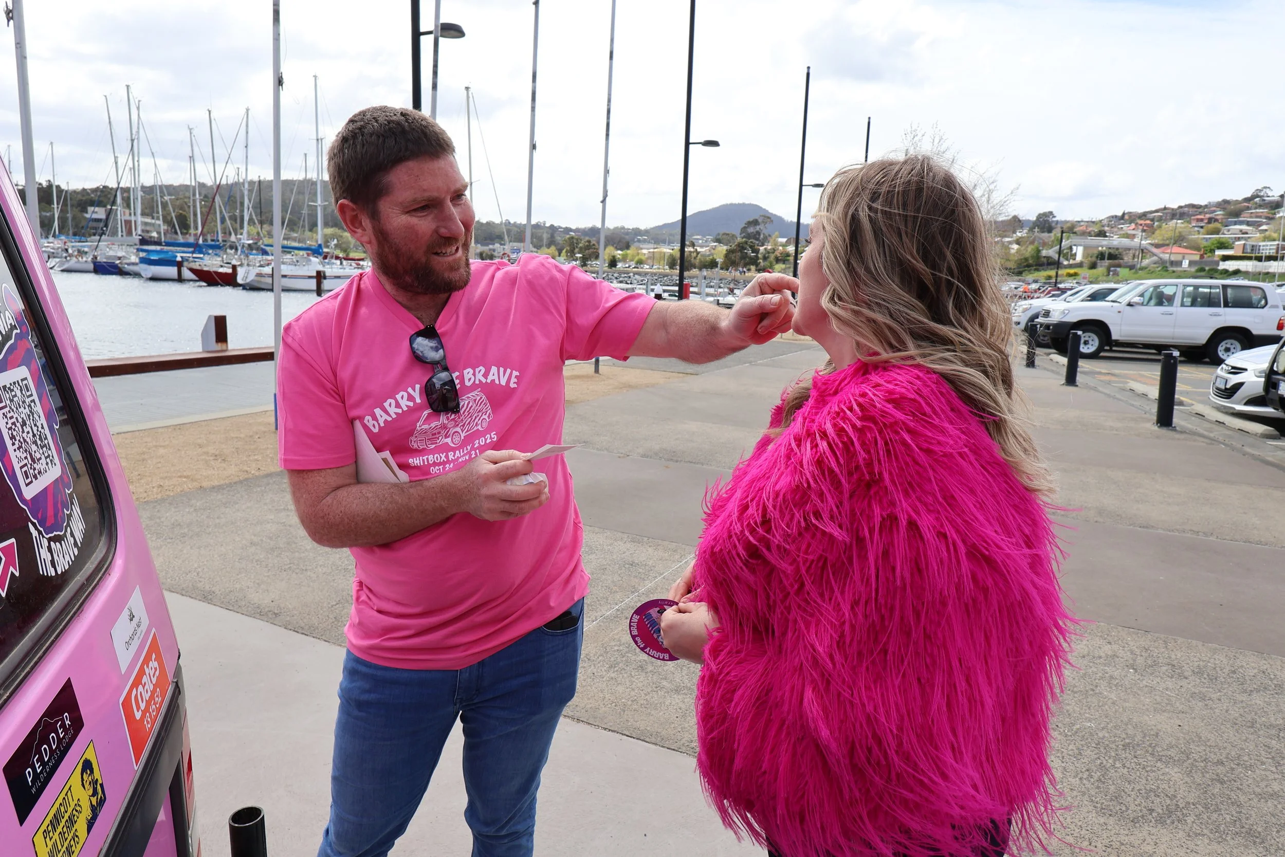 A man wearing a pink t-shirt and sunglasses is handing a sticker to a woman wearing a bright pink furry coat at a marina. There are sailboats in the water, parked cars, and hills in the background.