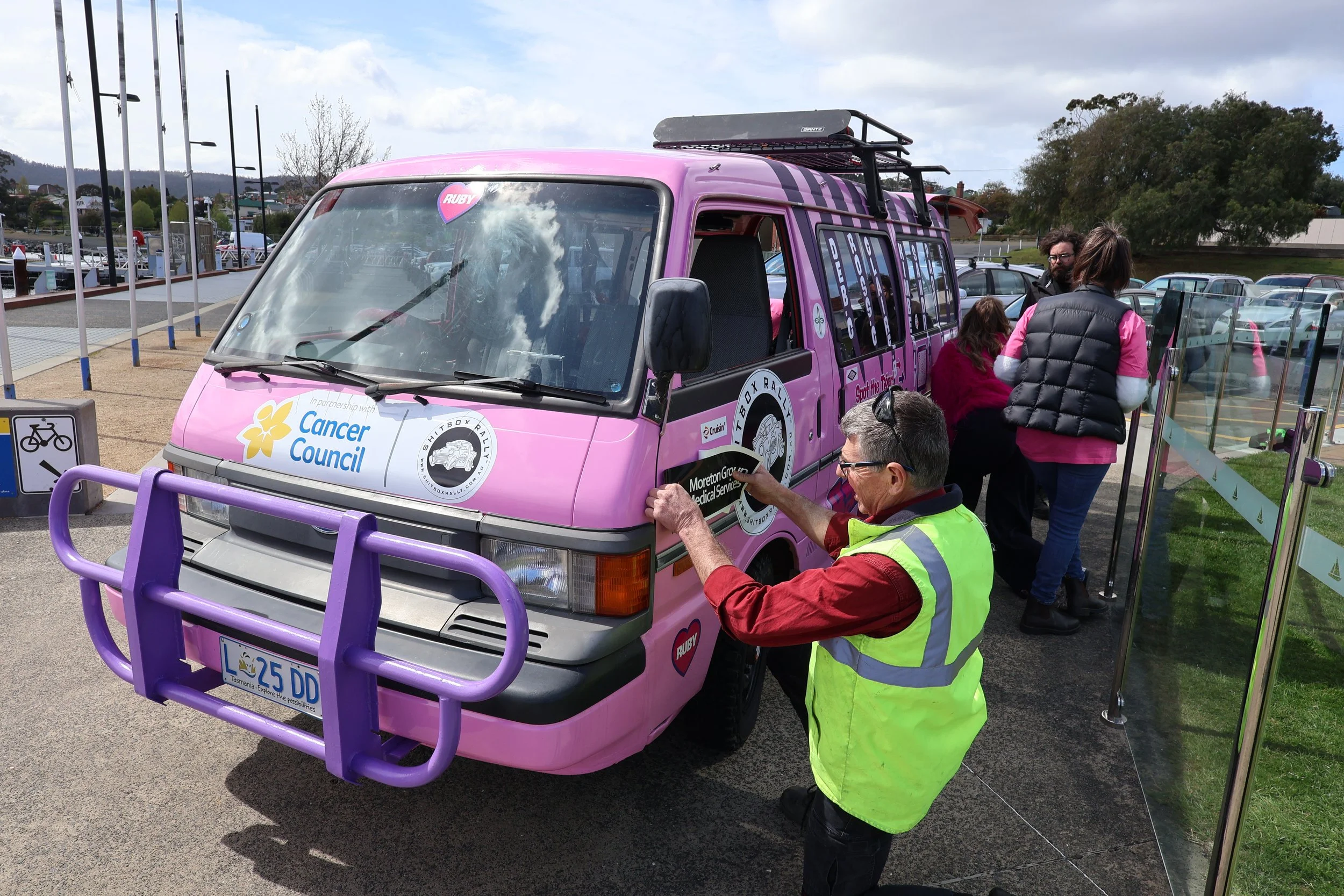A group of people, including a man with gray hair and a yellow safety vest, gather around a pink van with logos and stickers supporting the Cancer Council and other organizations. The van has a purple front bumper and several rooftop racks. They seem