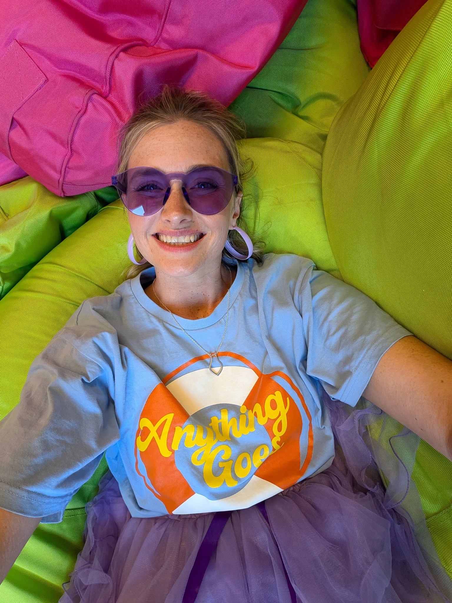 A woman lying on a colorful, multi-colored bed with pink, green, and purple sections, smiling and wearing sunglasses, a t-shirt with the words 'Anything Goes,' and a purple tulle skirt.