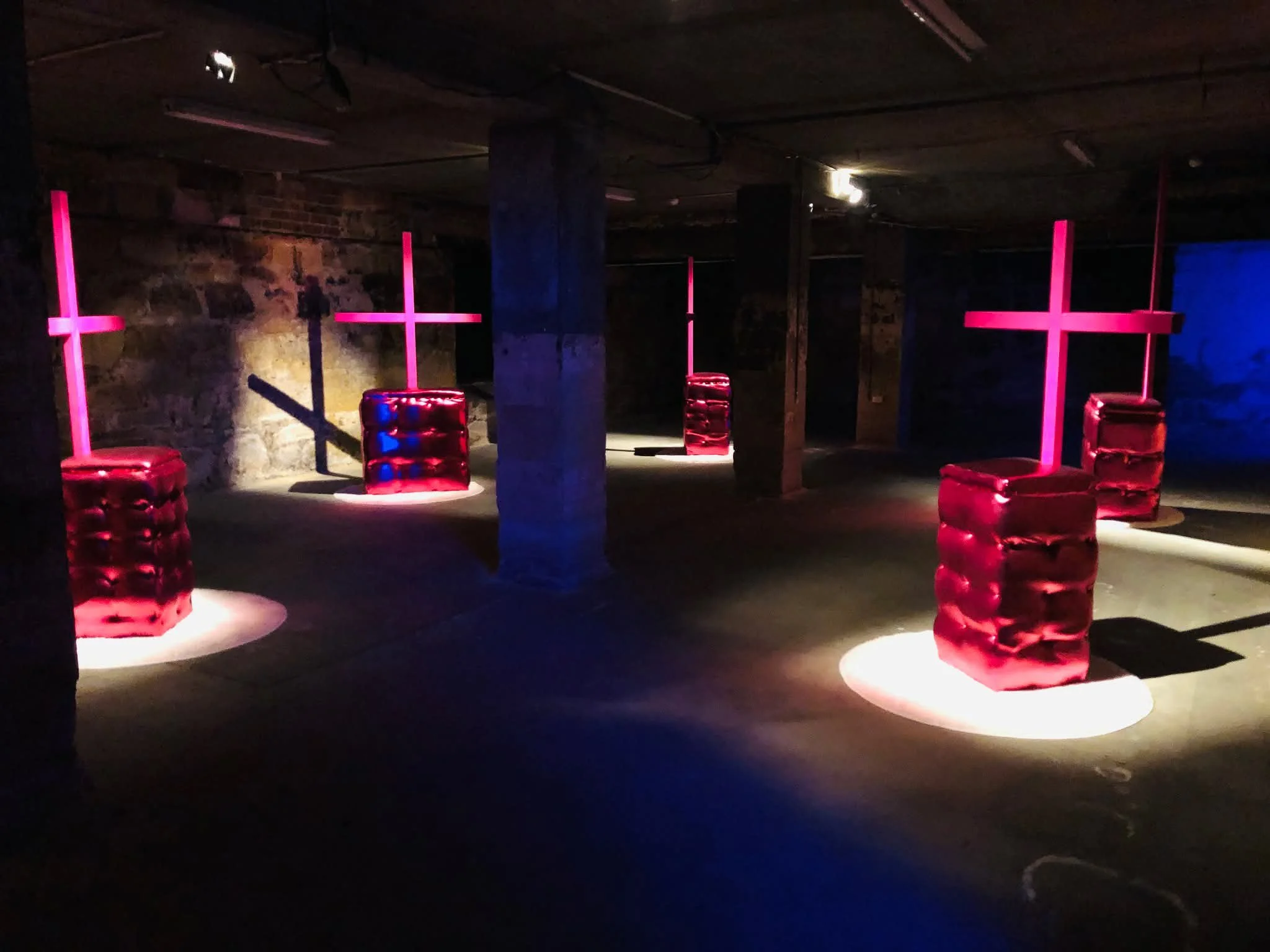 An underground space with brick walls and concrete support posts, illuminated by colorful lighting. There are five illuminated crosses with pink lighting, casting shadows on the floor. Red cushioned seats are placed near the crosses.