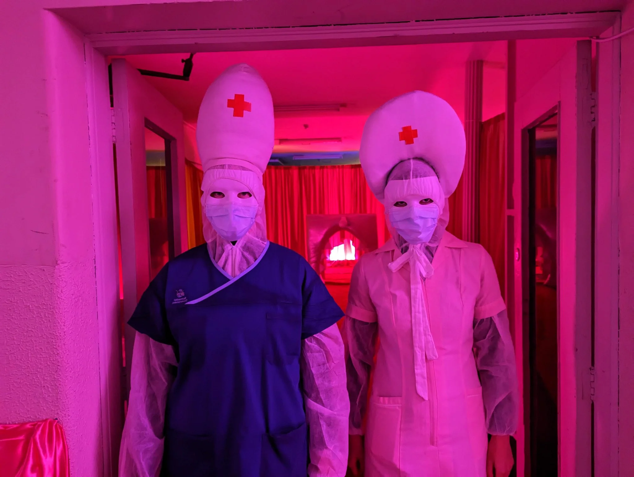 Two individuals dressed in healthcare attire with large nurse hats featuring red cross symbols, wearing face masks and standing under pink lighting in a corridor.