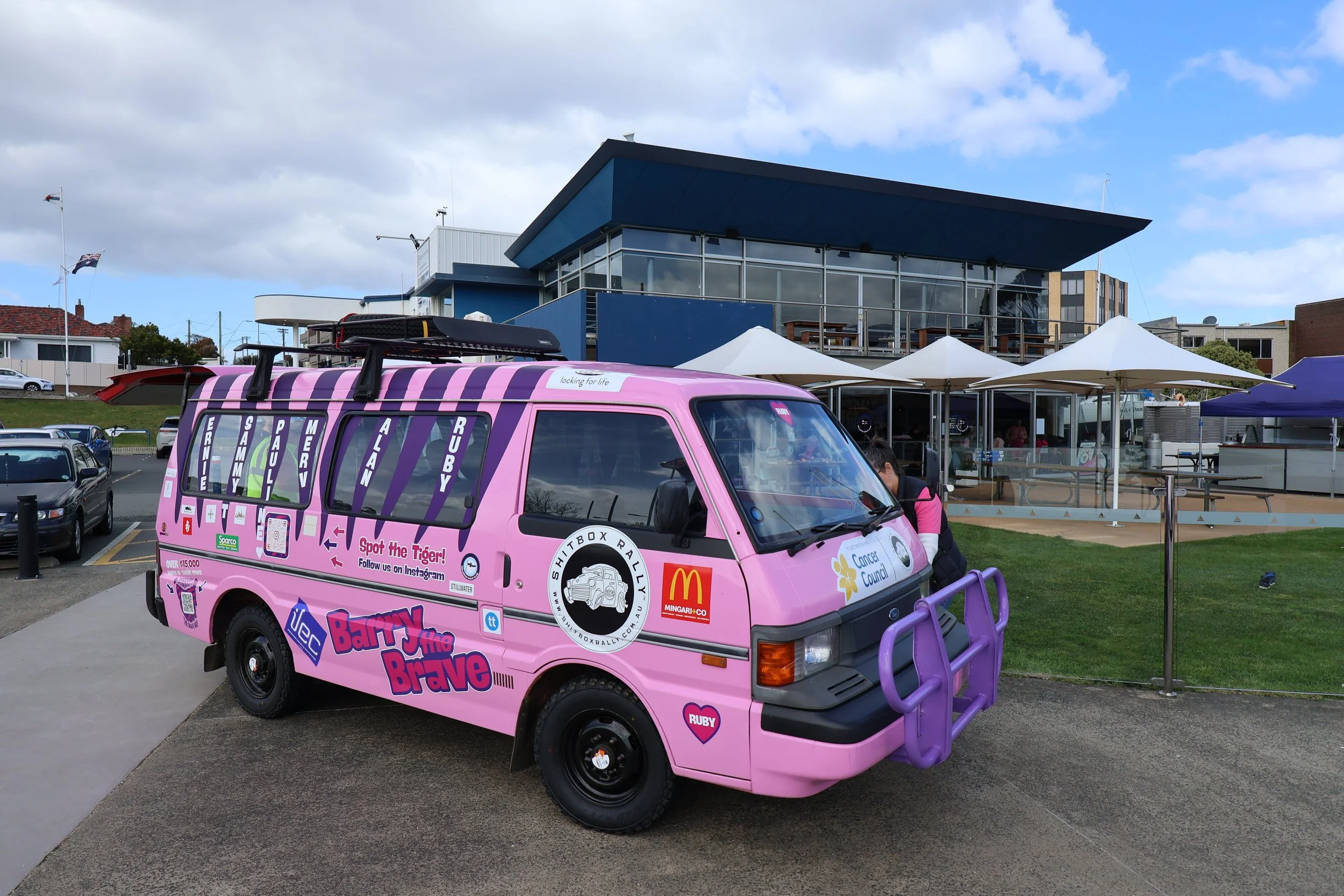 Pink van decorated with various stickers and slogans, parked in a lot outside a modern building with glass windows and outdoor seating. The van has logos and social media information, promoting an event or campaign.