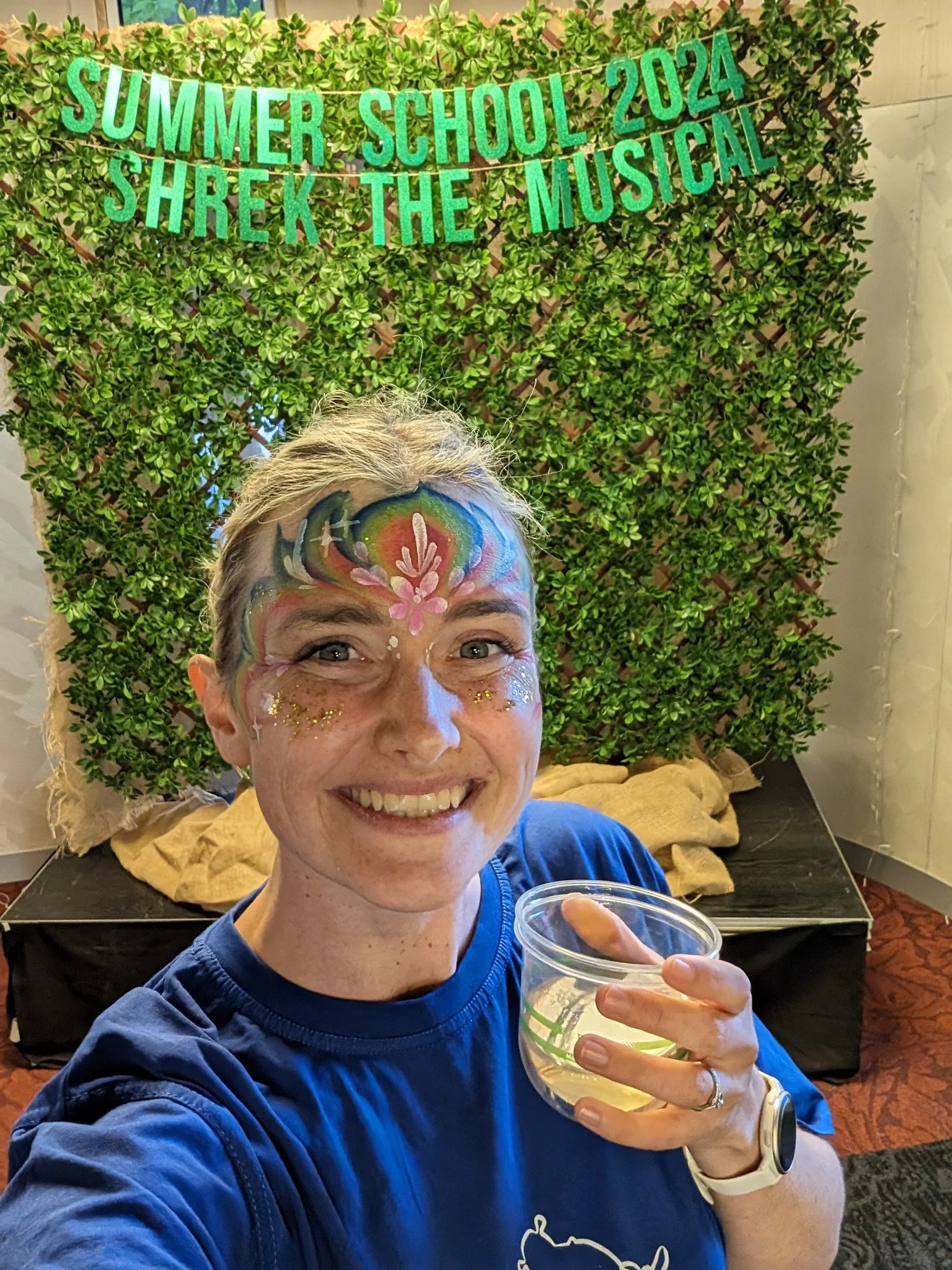 A woman with face paint of colorful flowers and sparkles, smiling and holding a cup of a yellow beverage, at a summer event with a green hedge backdrop and a sign reading 'Summer School 2024 Shrek the Musical' in green letters.